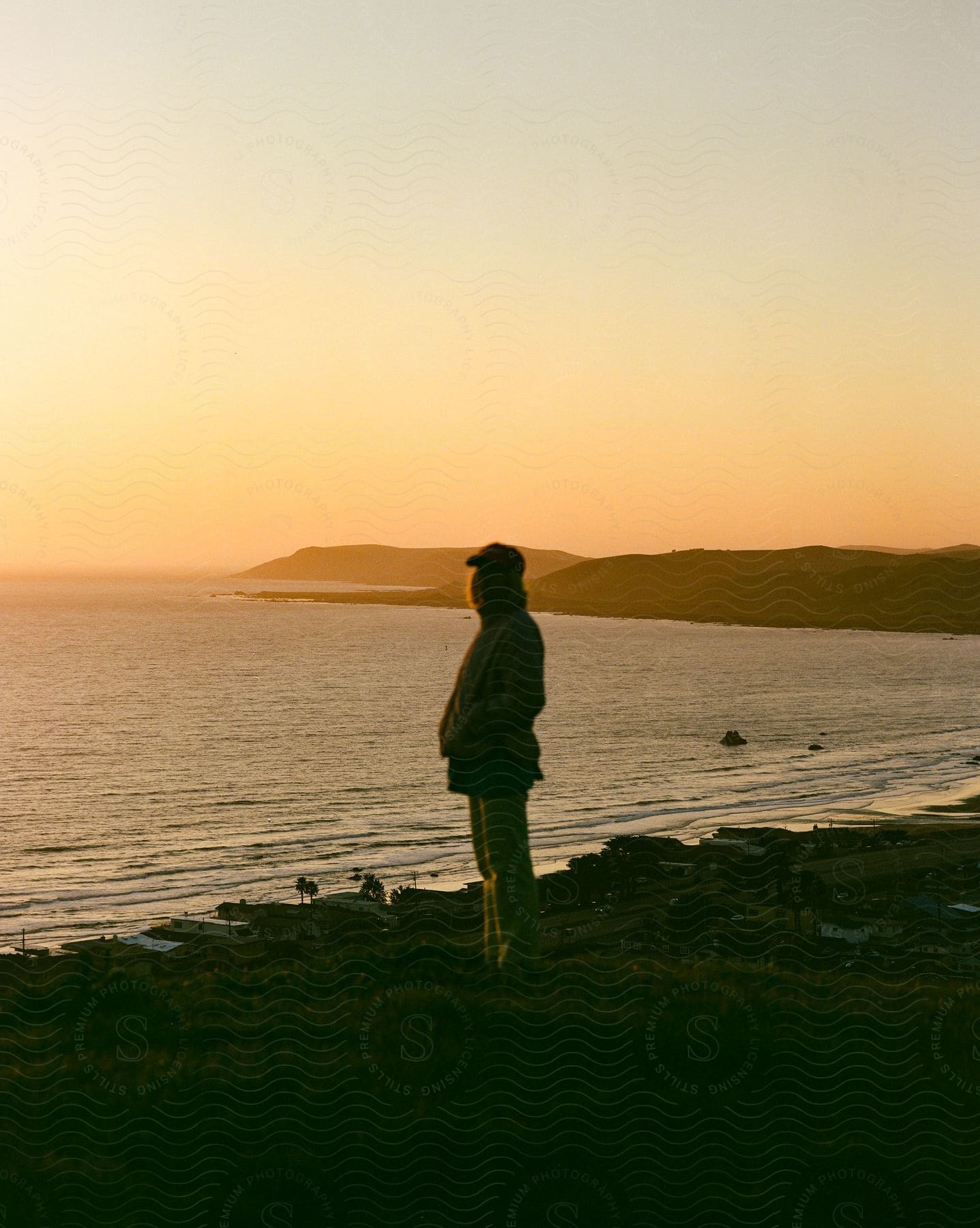 A person in a baseball cap stands on a grassy hill above a coastal town as the sun sets by the ocean