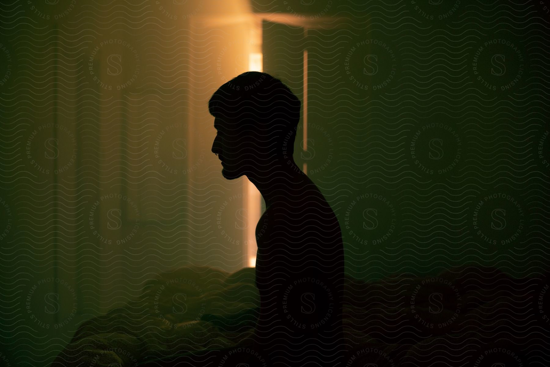 A man sits up in bed in a dark bedroom silhouetted against the light from the doorway