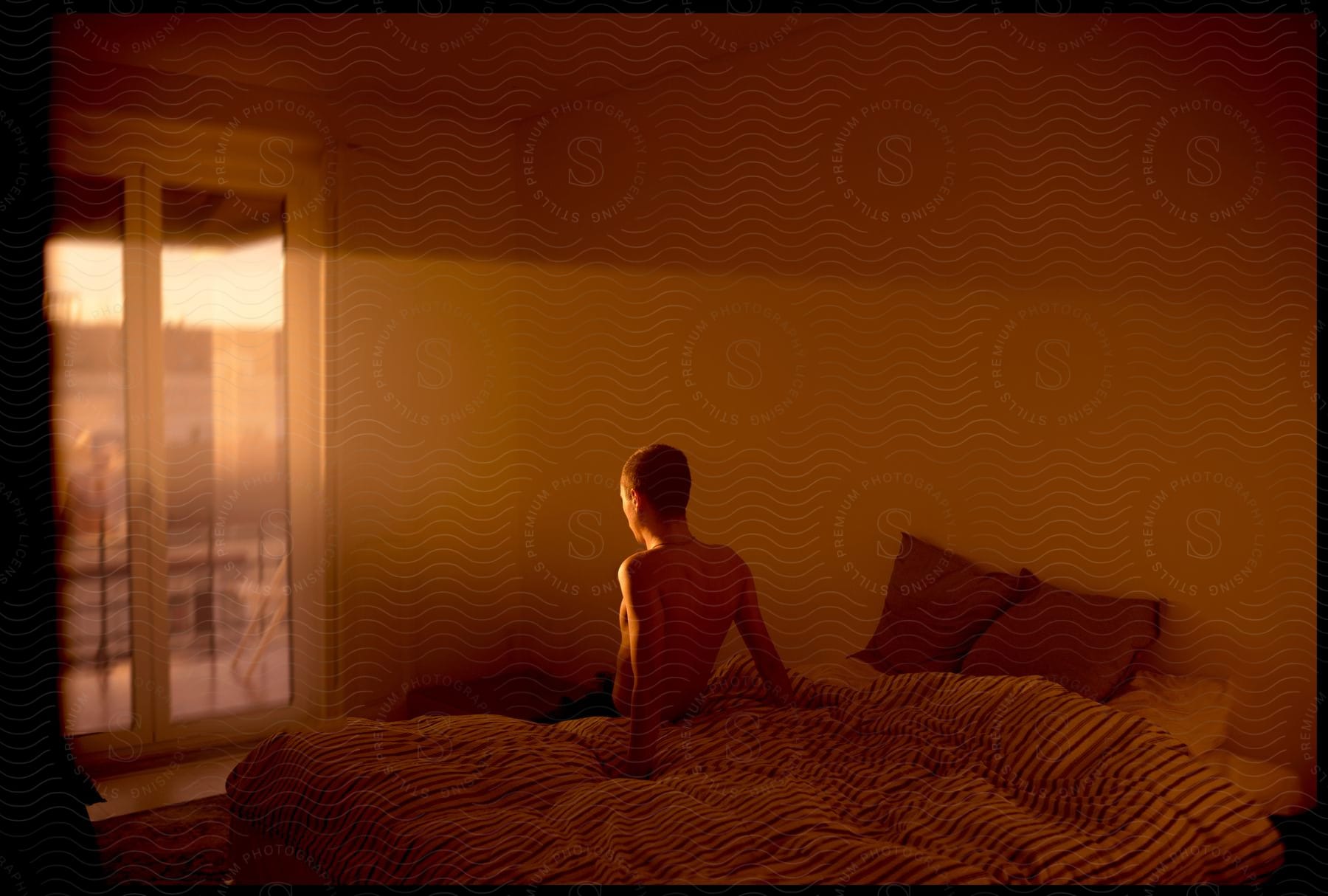 A man sitting on a bed in his bedroom looking at the sunrise through the balcony doors