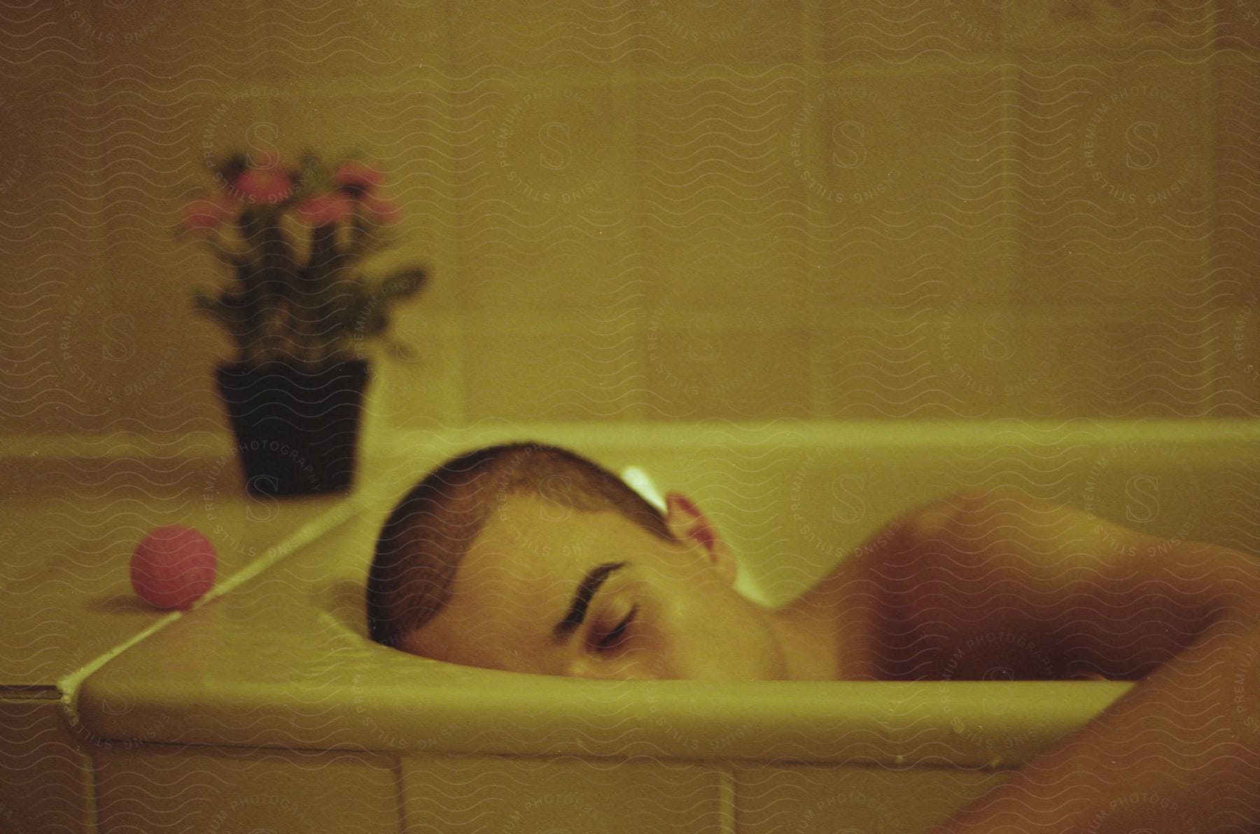A man lying on his side in a bathtub with his arm over the side a ball and a plant nearby