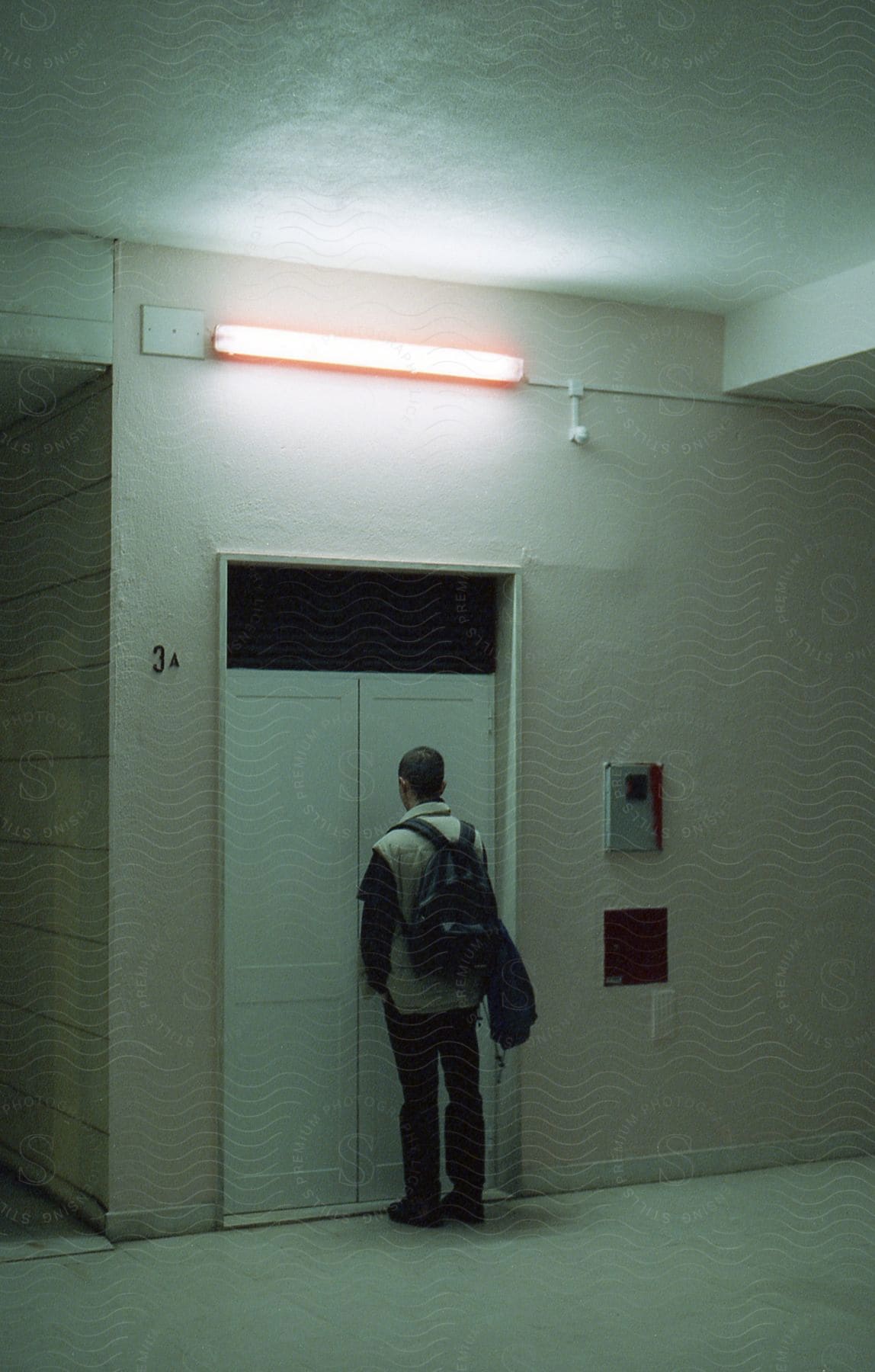 A man with a backpack stands by a closed door illuminated by a fluorescent light
