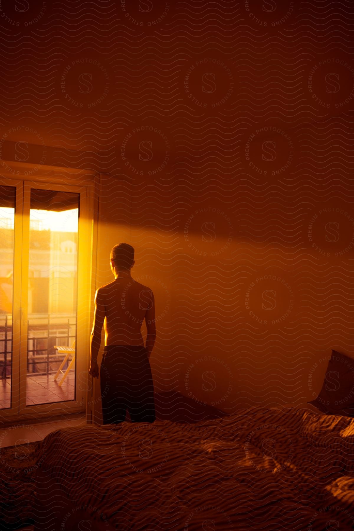 A man standing shirtless in a bedroom observing the sunrise through the balcony doors