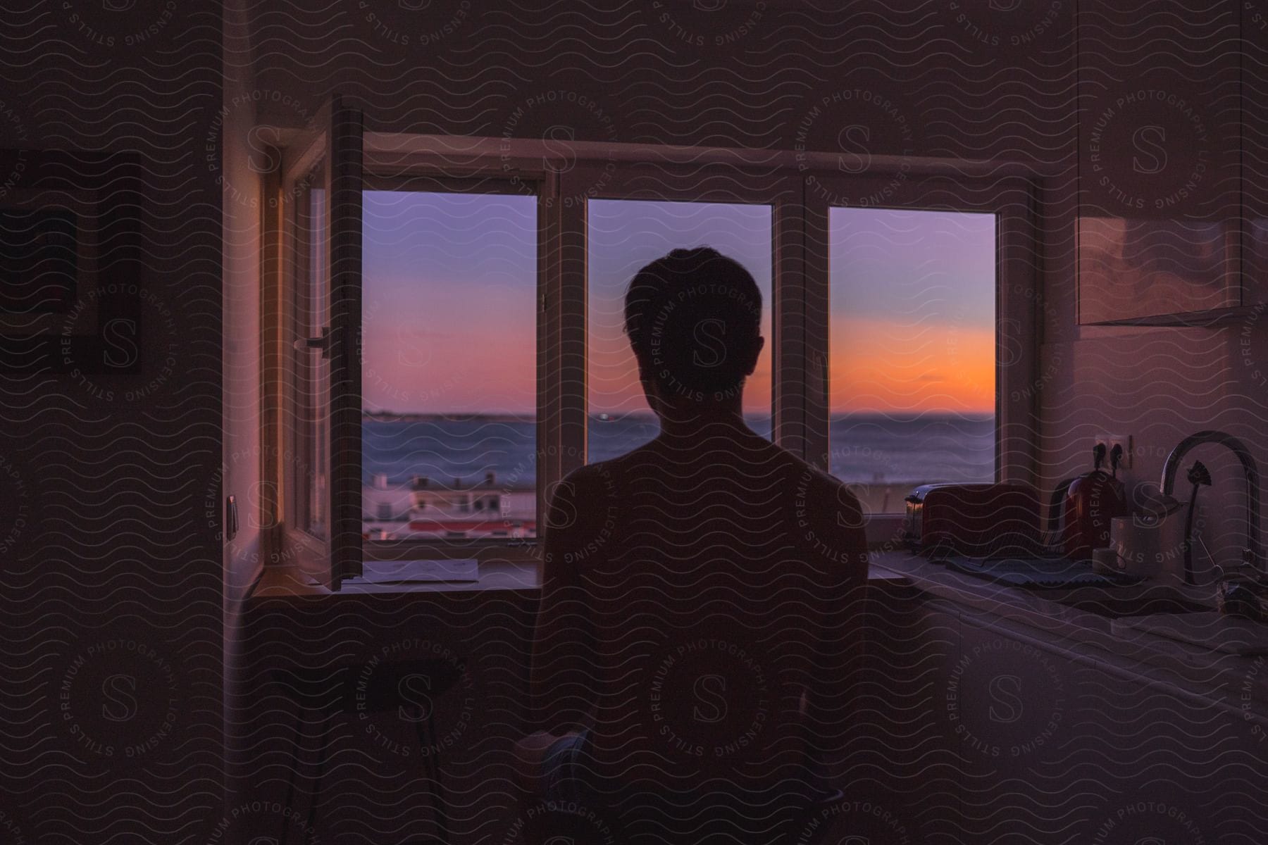 A man sitting by a kitchen window watching the sunset over the ocean