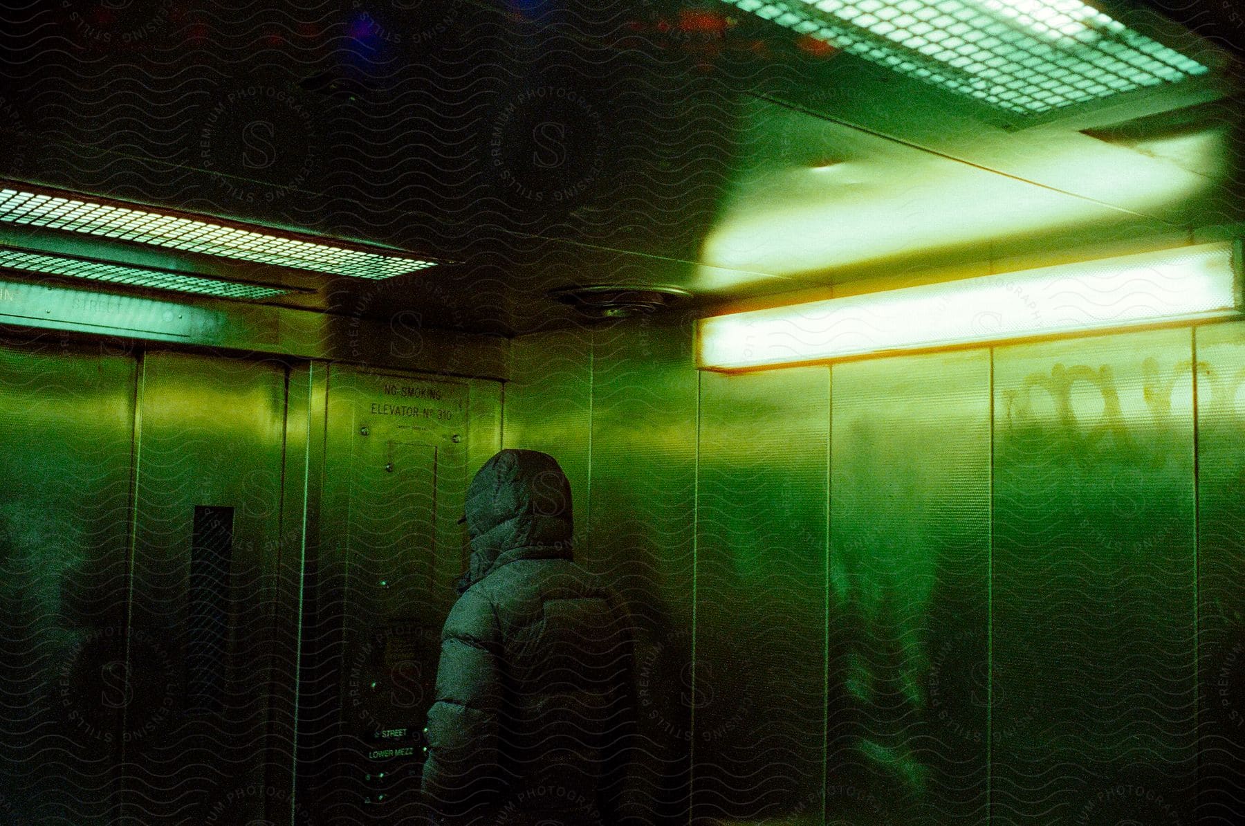 A person wearing a hooded jacket is riding a welllit elevator