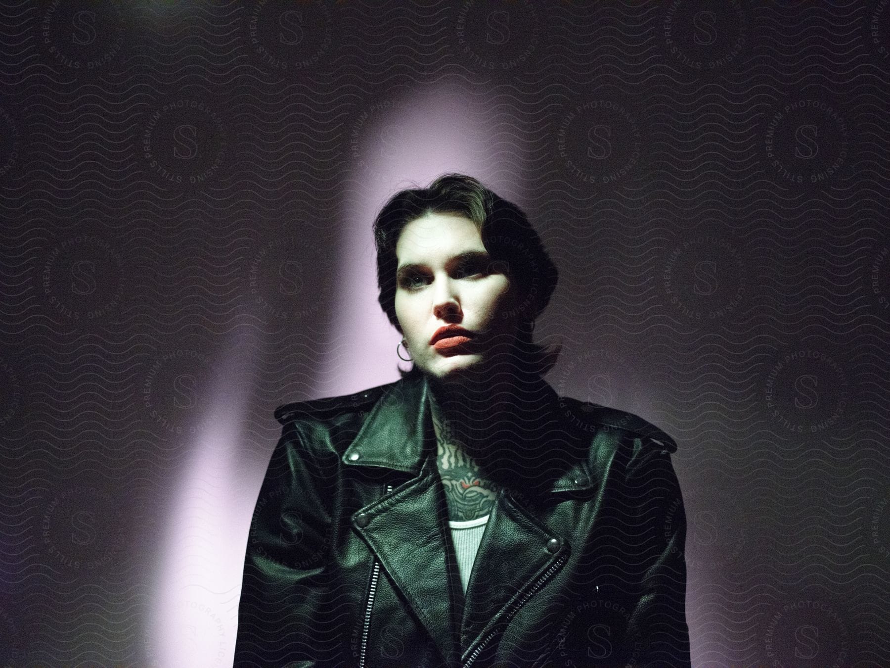 In a dark room with purple walls a young lady stands under focused light wearing a black leather jacket red lipstick and a chest tattoo