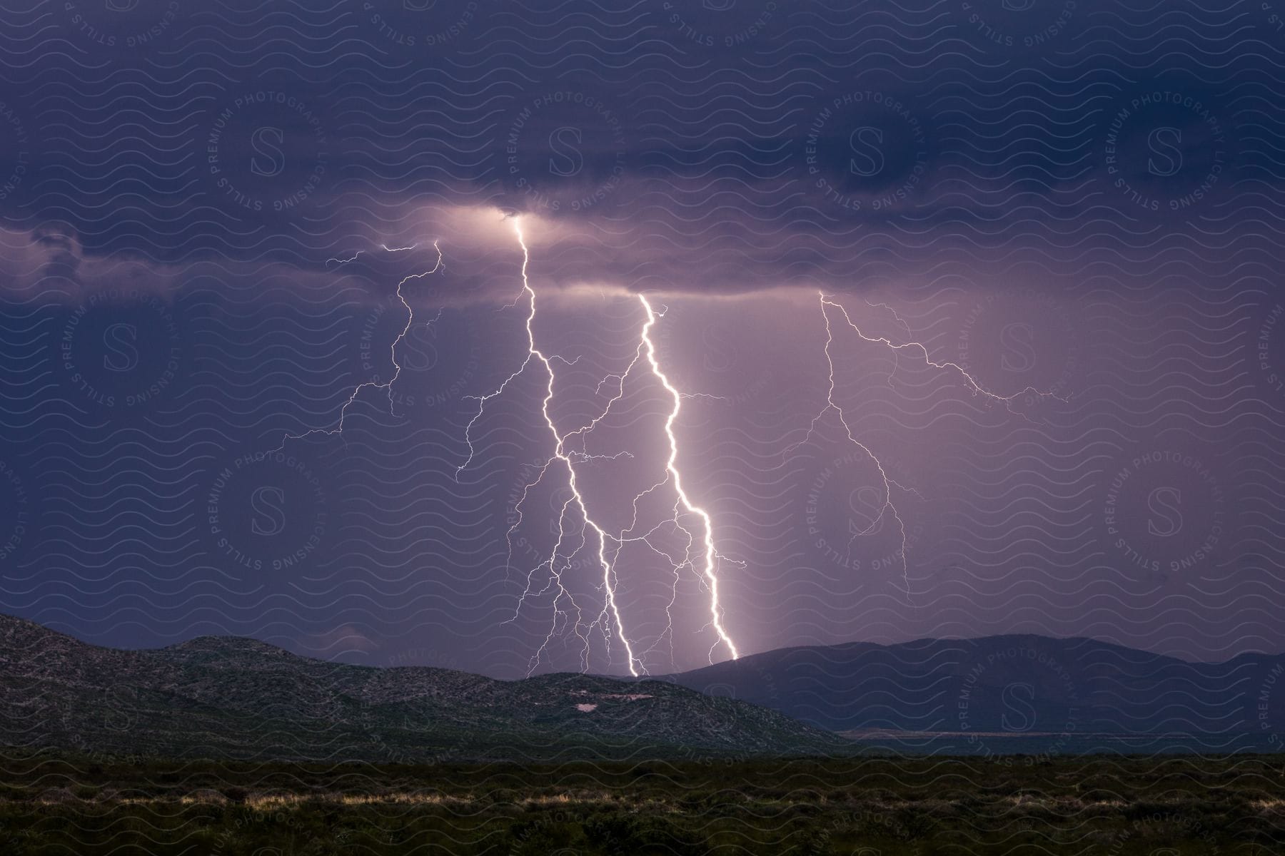 Multiple lightning bolts strike a mountain range illuminating the grasslands in the foreground