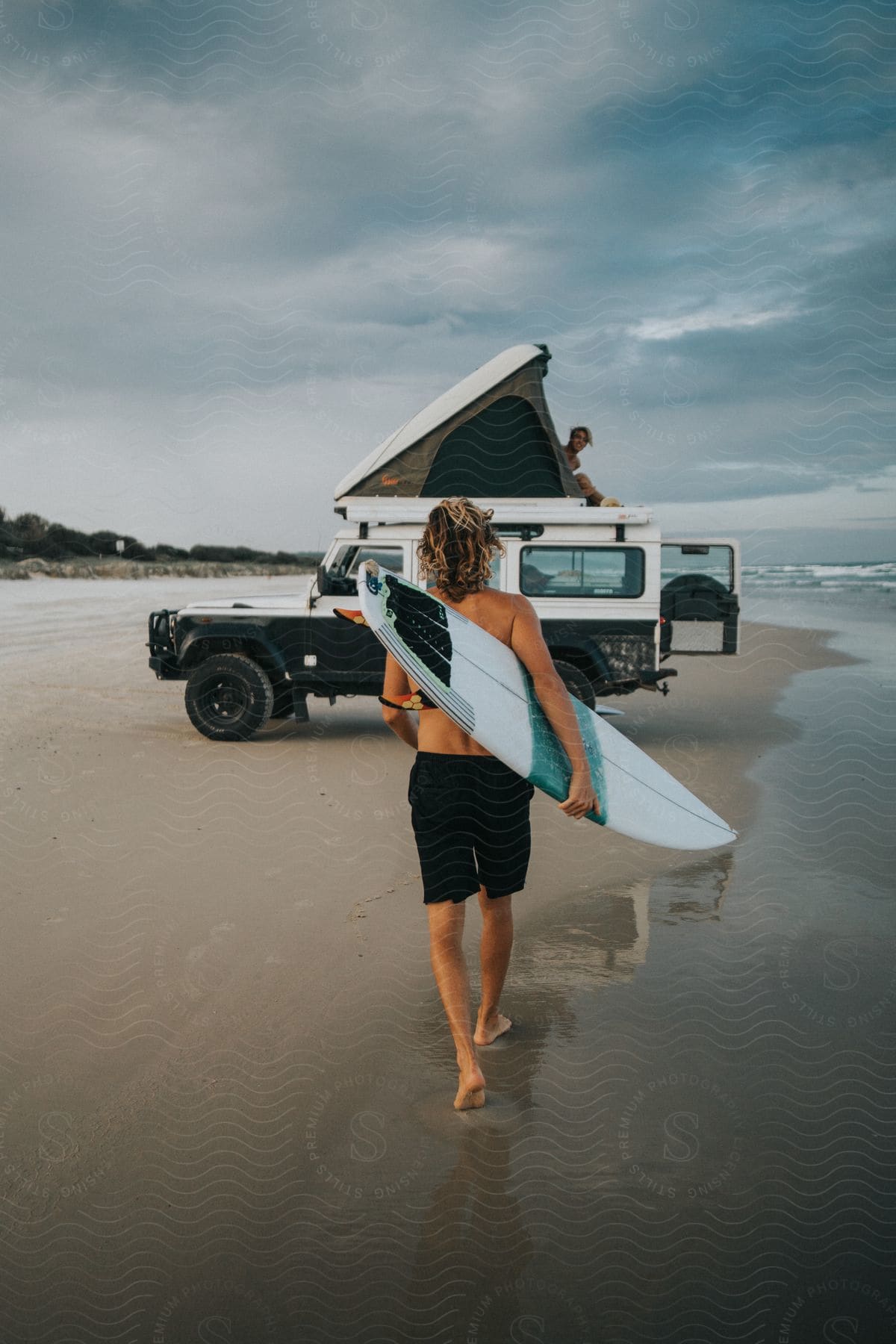 A person with a surfboard walking towards a jeep near the water