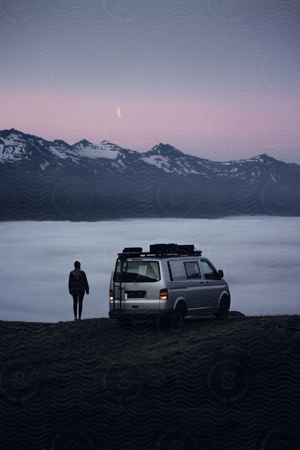 A woman in winter clothing stands next to a van on a hill overlooking a moonlit mountain range and a foggy valley