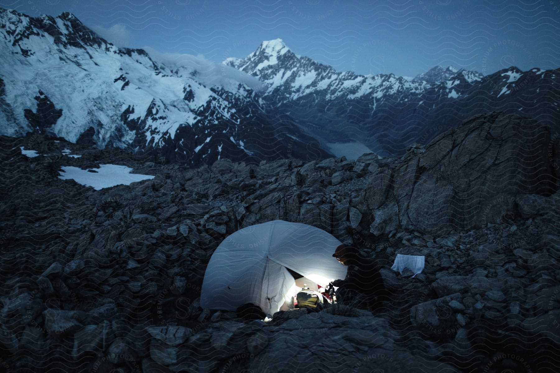 A Man Sits Outside A Tent In Rocky Terrain With Snowy Mountains In The Background At Night Time