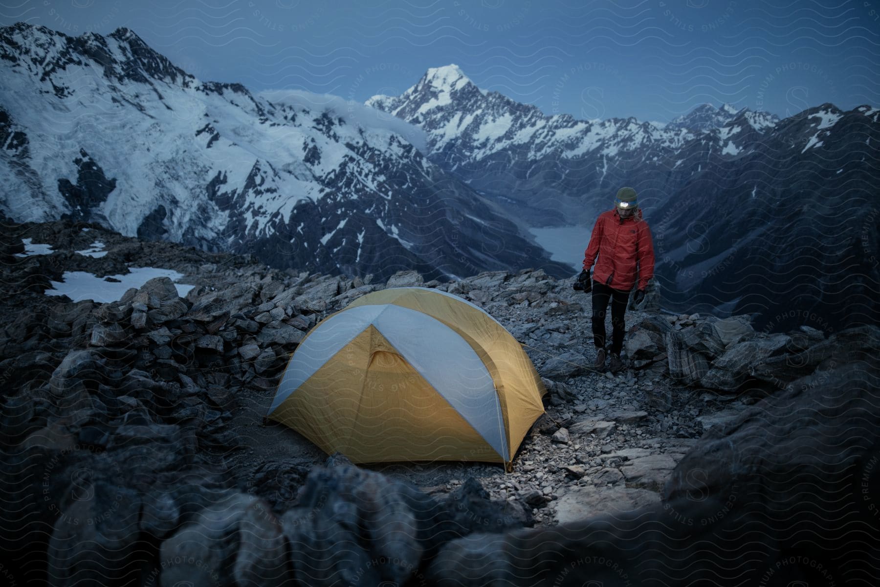 A hiker in an orange jacket stands next to a tent on a mountain overlooking a valley with a lake and snowcapped peaks in the background