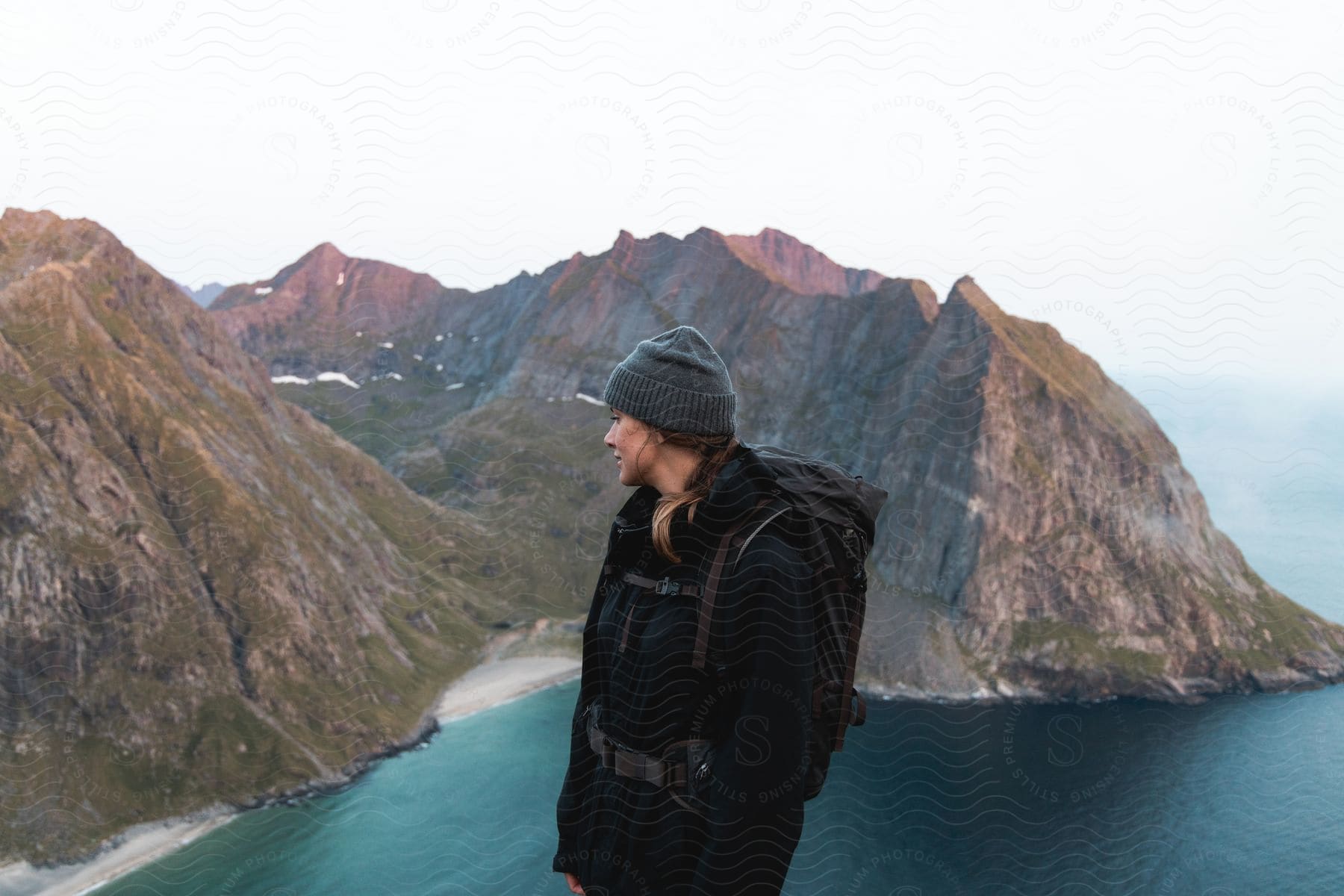 A woman in winter clothing stands on a mountain looking at a mountain range that extends into the ocean