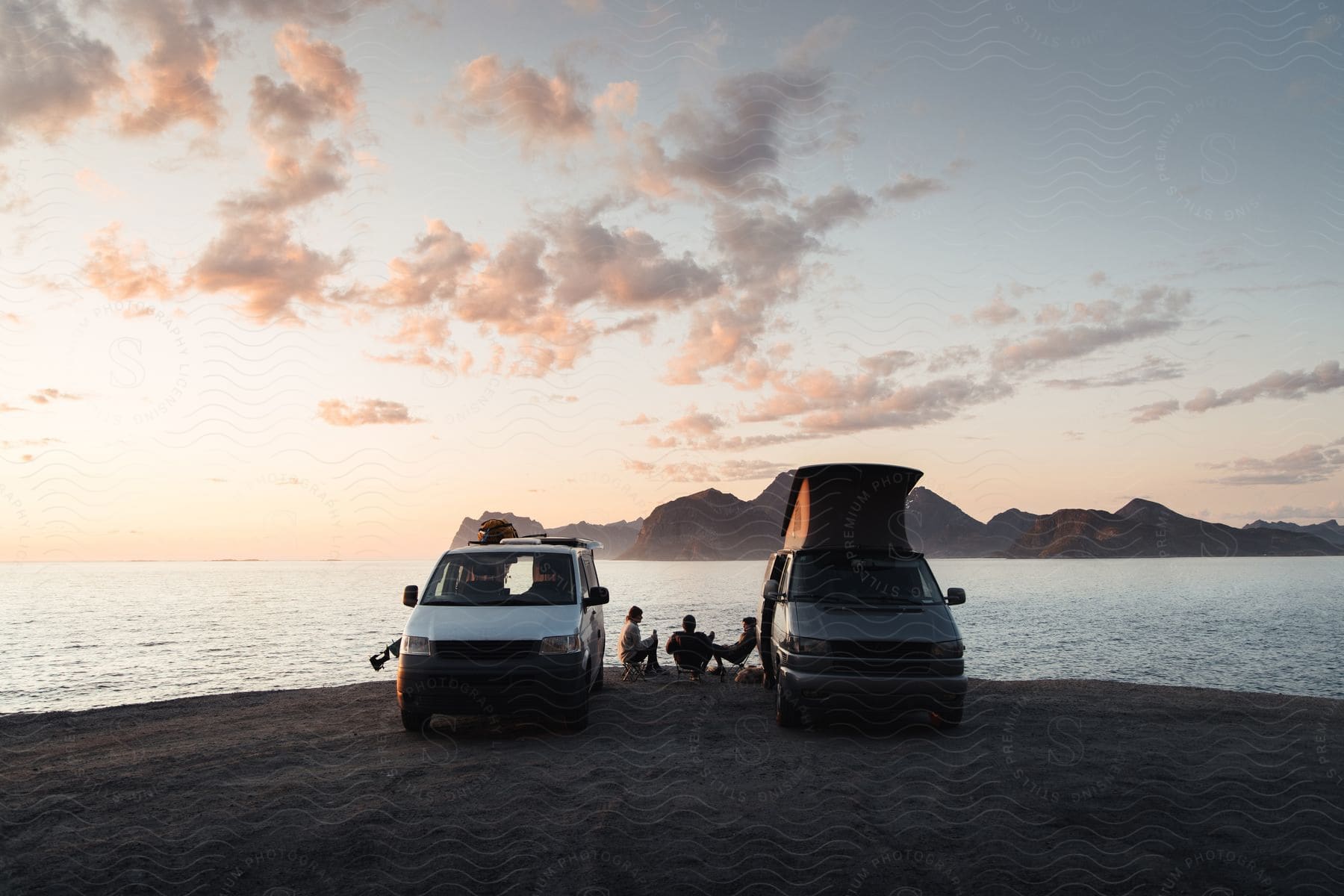 Two vans parked on a beach at sunset with people sitting in a circle on the sand a mountain island in the distance and cloud puffs in the sky