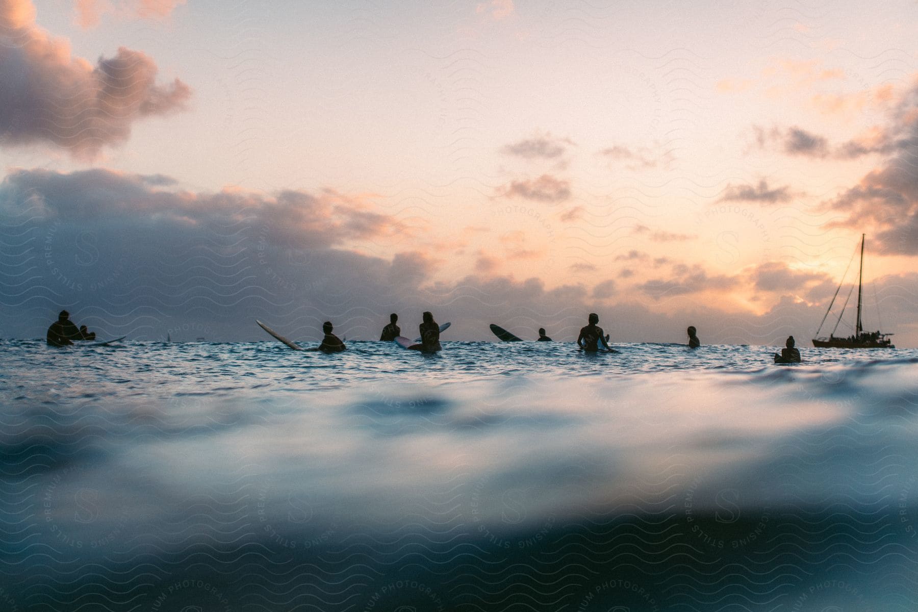 People riding surfboards in the ocean at sunset with a sailboat in the background