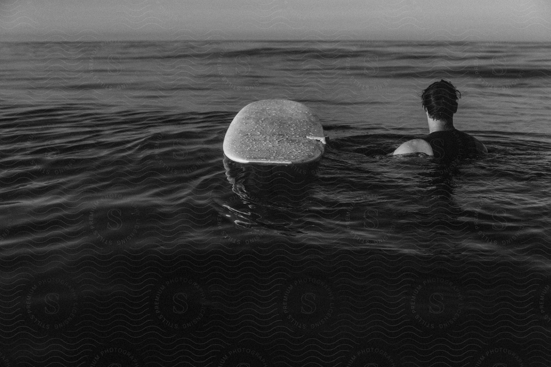 A man in the ocean wading in water next to a surfboard looking out over the ocean