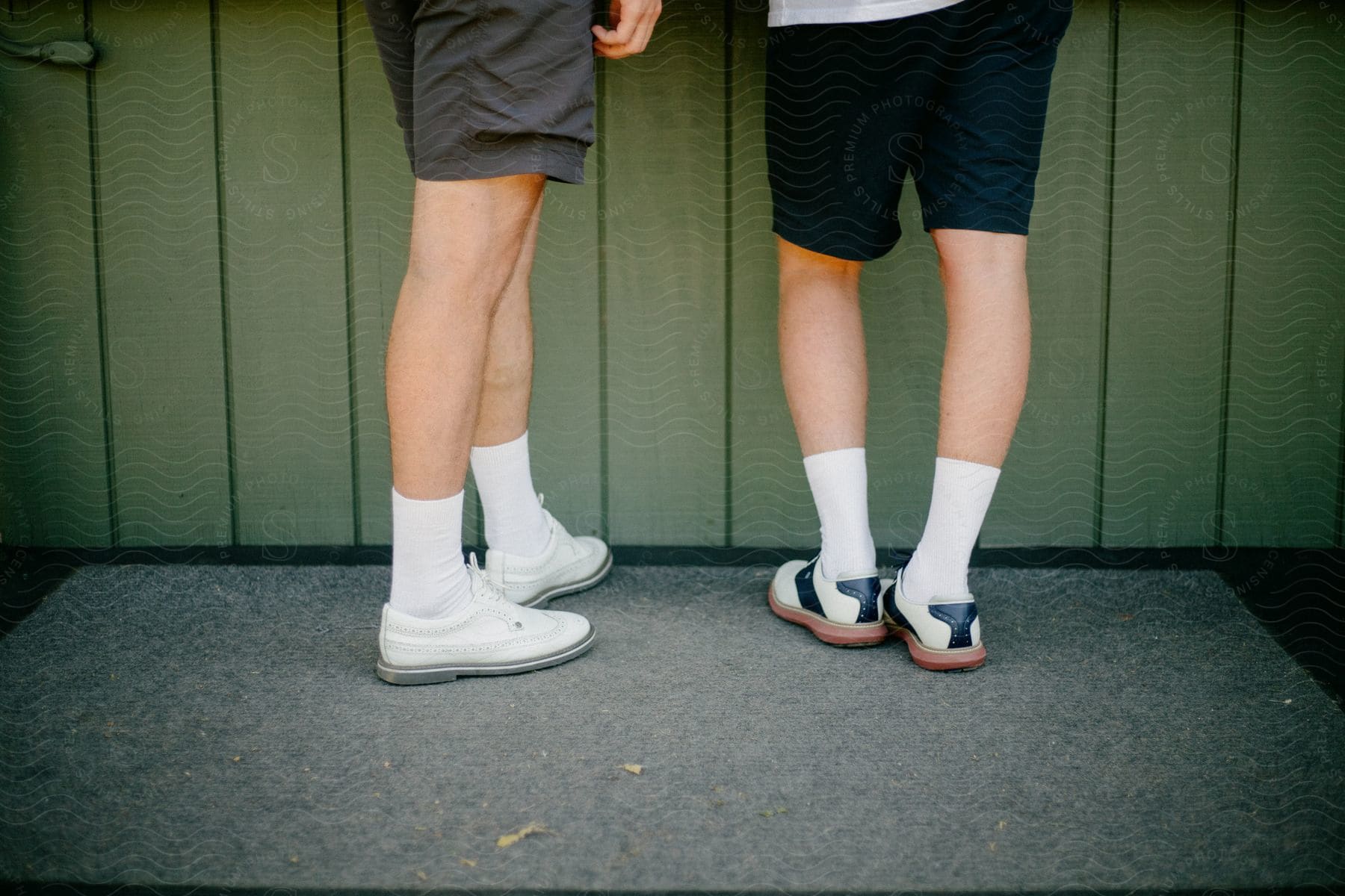 Two mens legs and feet in golf shoes standing on a concrete slab in front of a green building