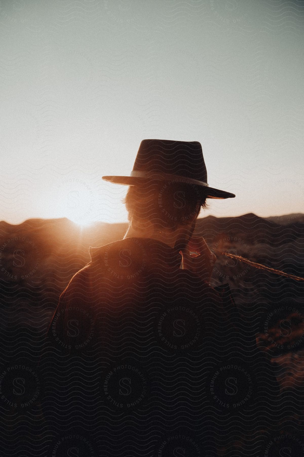 A man wearing a cowboy hat is standing in the desert looking toward mountains as the setting sun shines