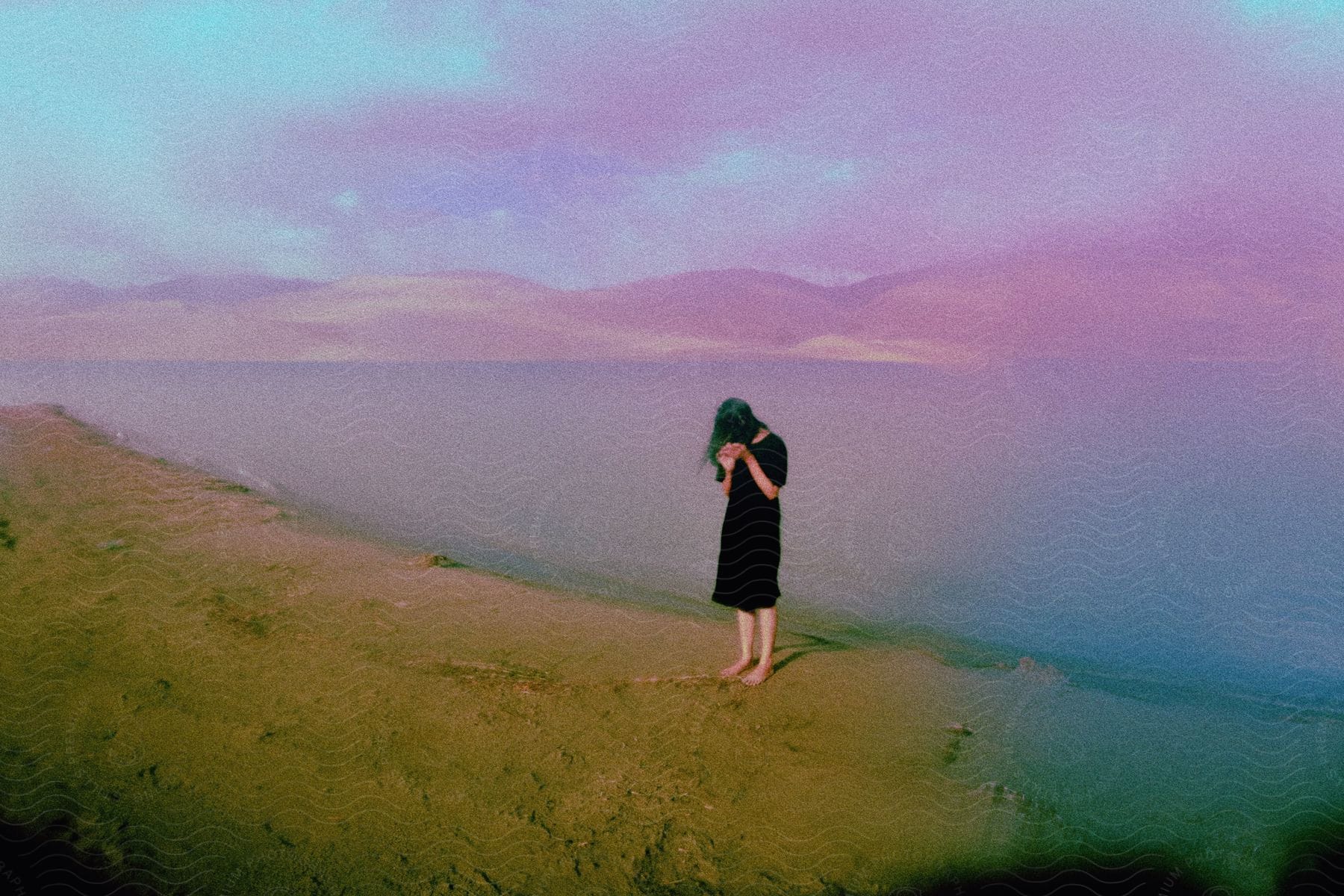 A teenage girl wearing a black dress is standing barefoot on the beach with mountains in the distance