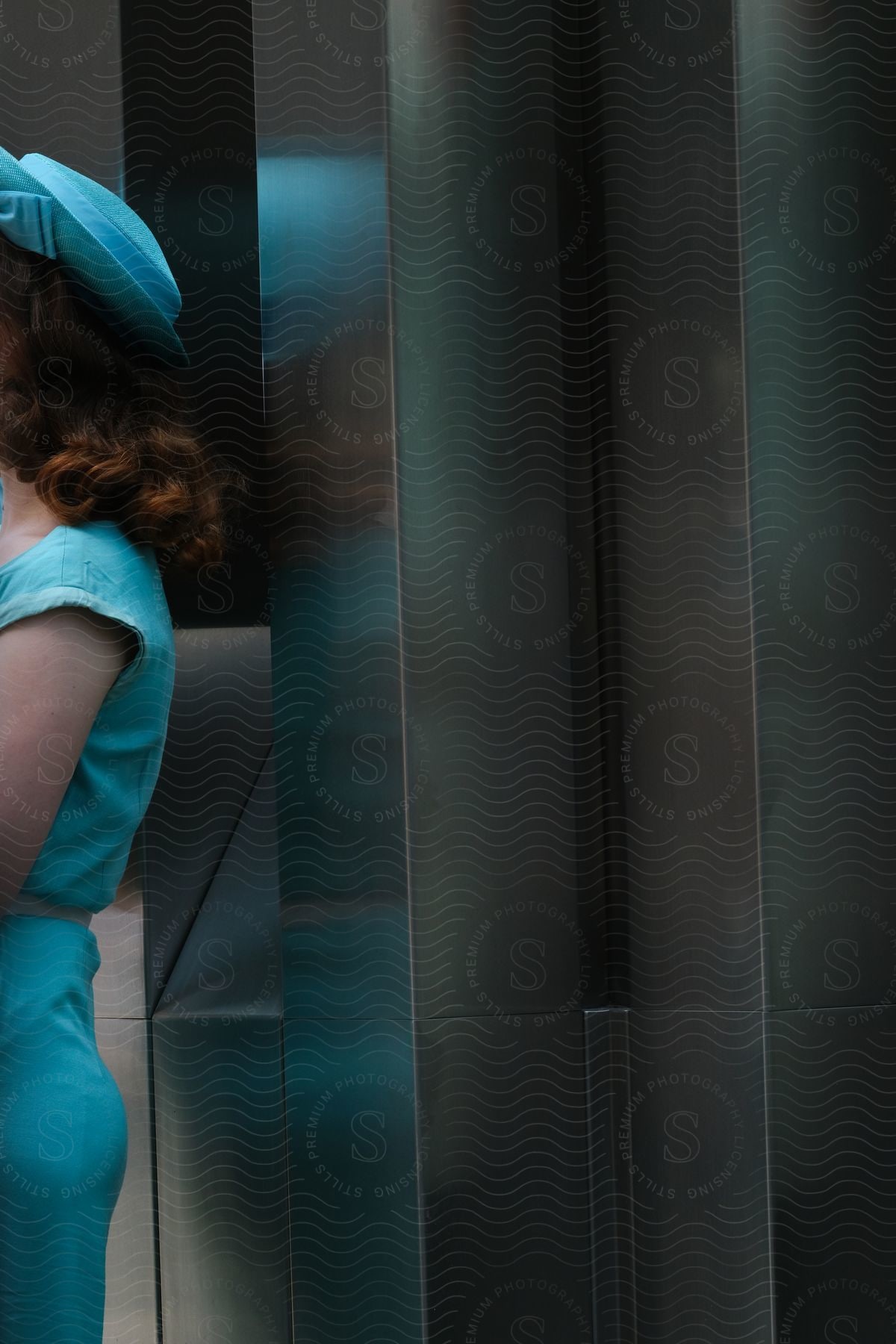 Redhead wearing a blue dress with a matching hat stands near metal posts in building lobby.