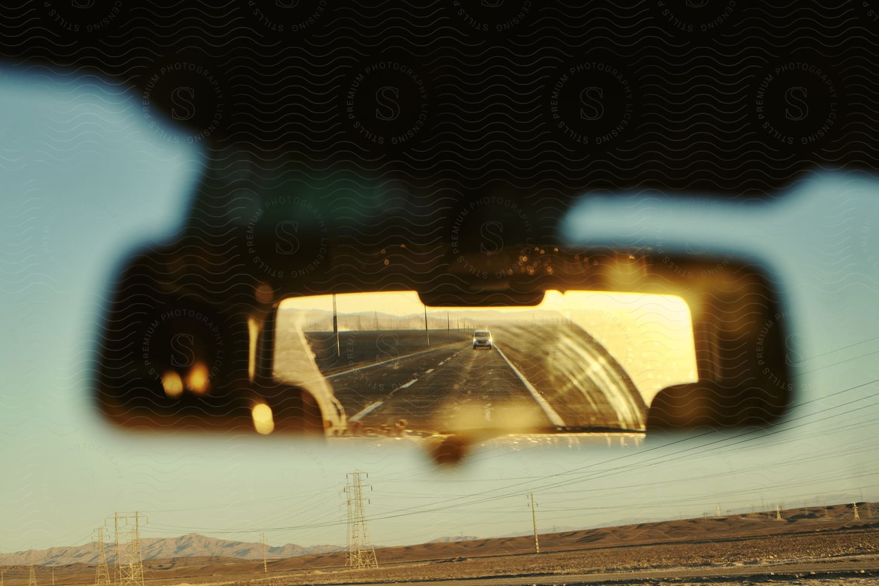 A car's rearview mirror reflects the sunset over a road in the desert, with the car's reflection visible in the mirror's reflection.