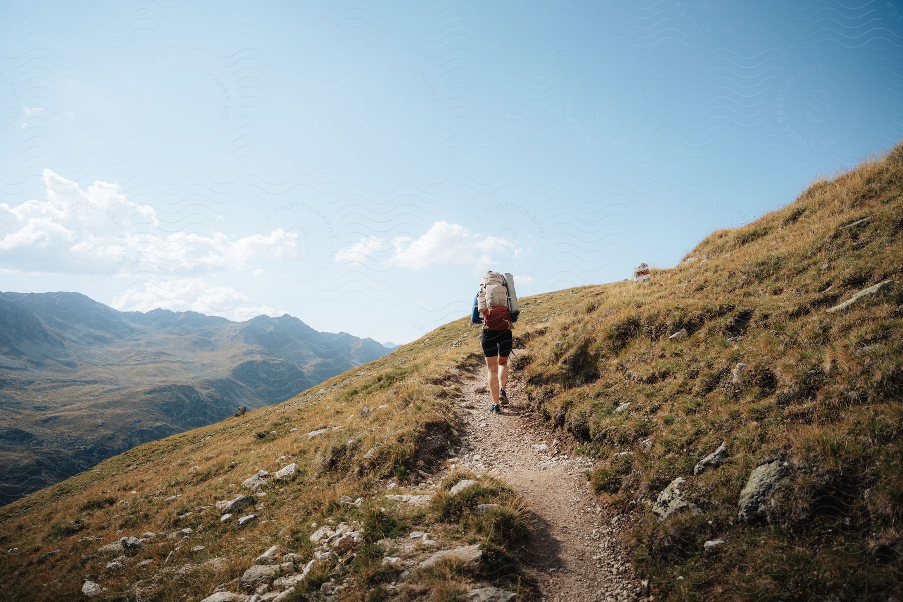 Stock photo of a man carrying a camping bag and dressed in shorts walks along a path leading up to the mountain. green grasses surround the path on both the left and right sides.