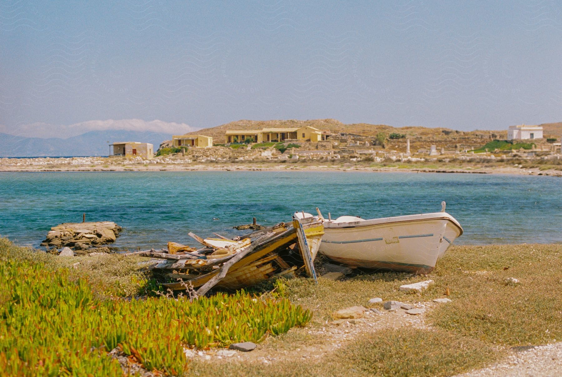 A boat sits on the shore, next to a destroyed one, overlooking the ocean and buildings in the distance.