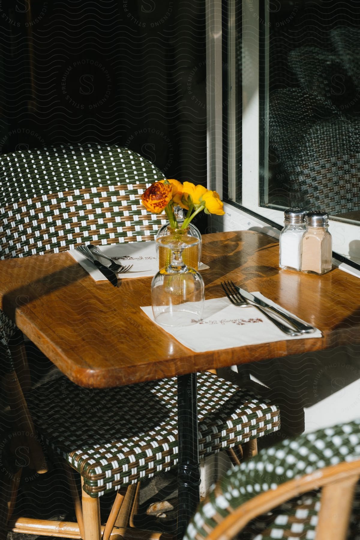 A table set for two in a restaurant with utensils, salt and pepper, and yellow flowers as a center piece.