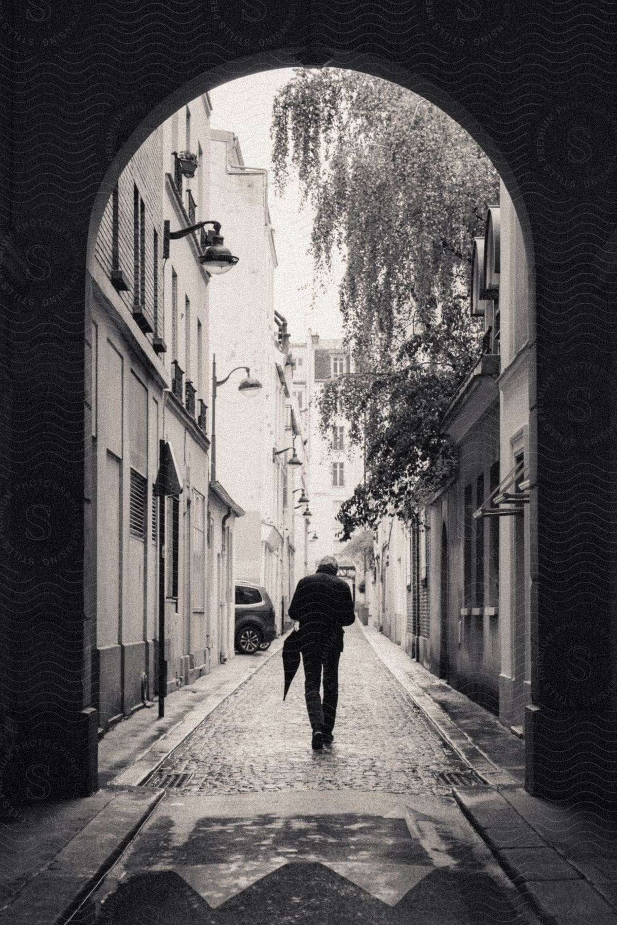 A man walks into the alley, holding an umbrella beside him, while a car is parked on the side. A tree provides cover to a building on the right, and the ground is still wet from a recent rainfall