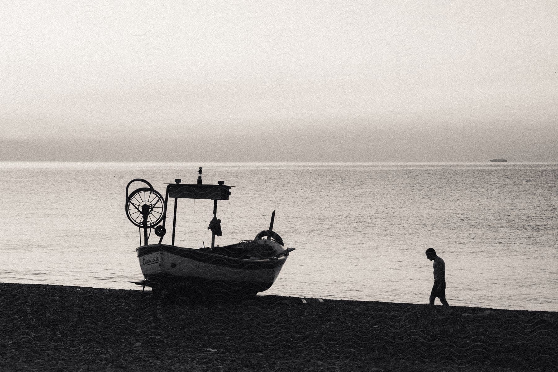 A man walks towards a boat docked on the beach, with a wheel in front, while the beautiful sea view serves as a scenic backdrop