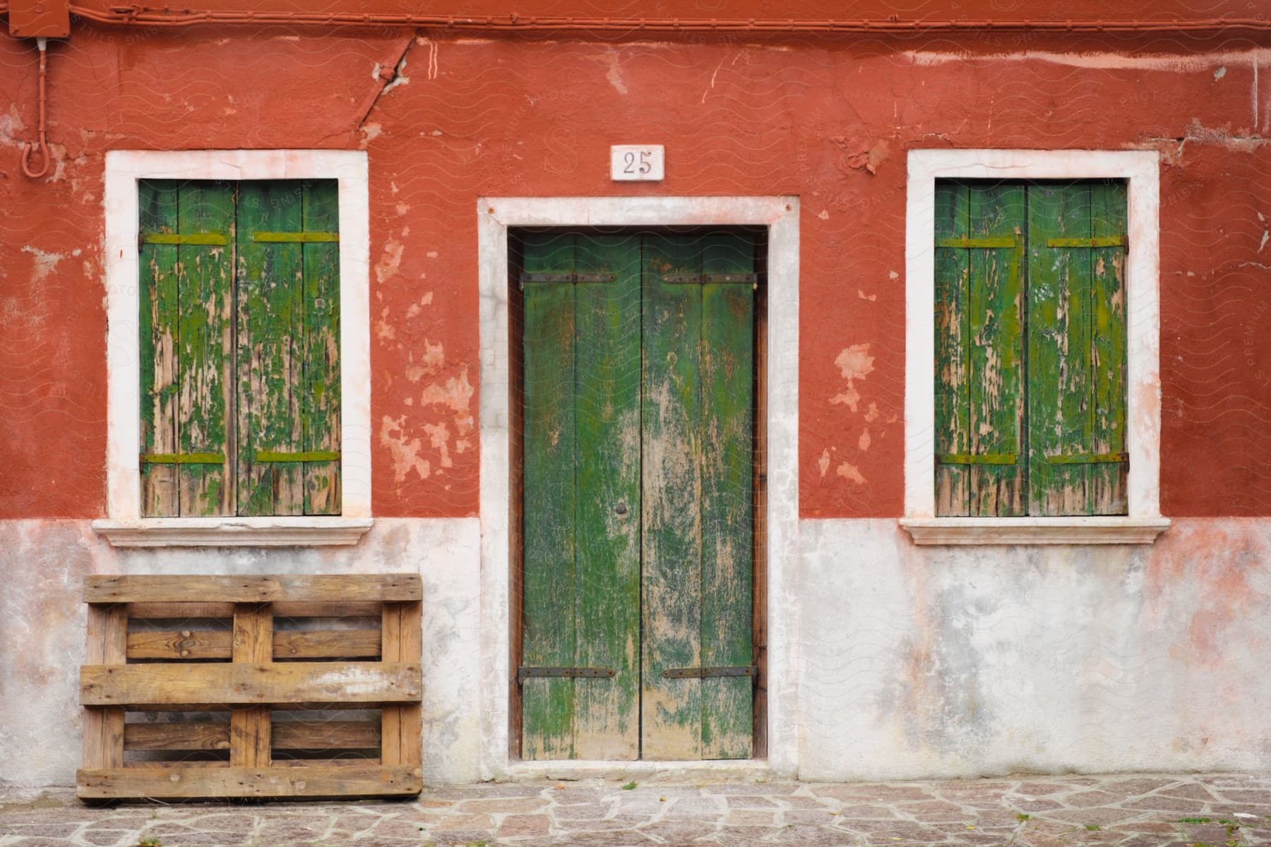 A wooden pallet leaning against a red house with worn green door and window shutters