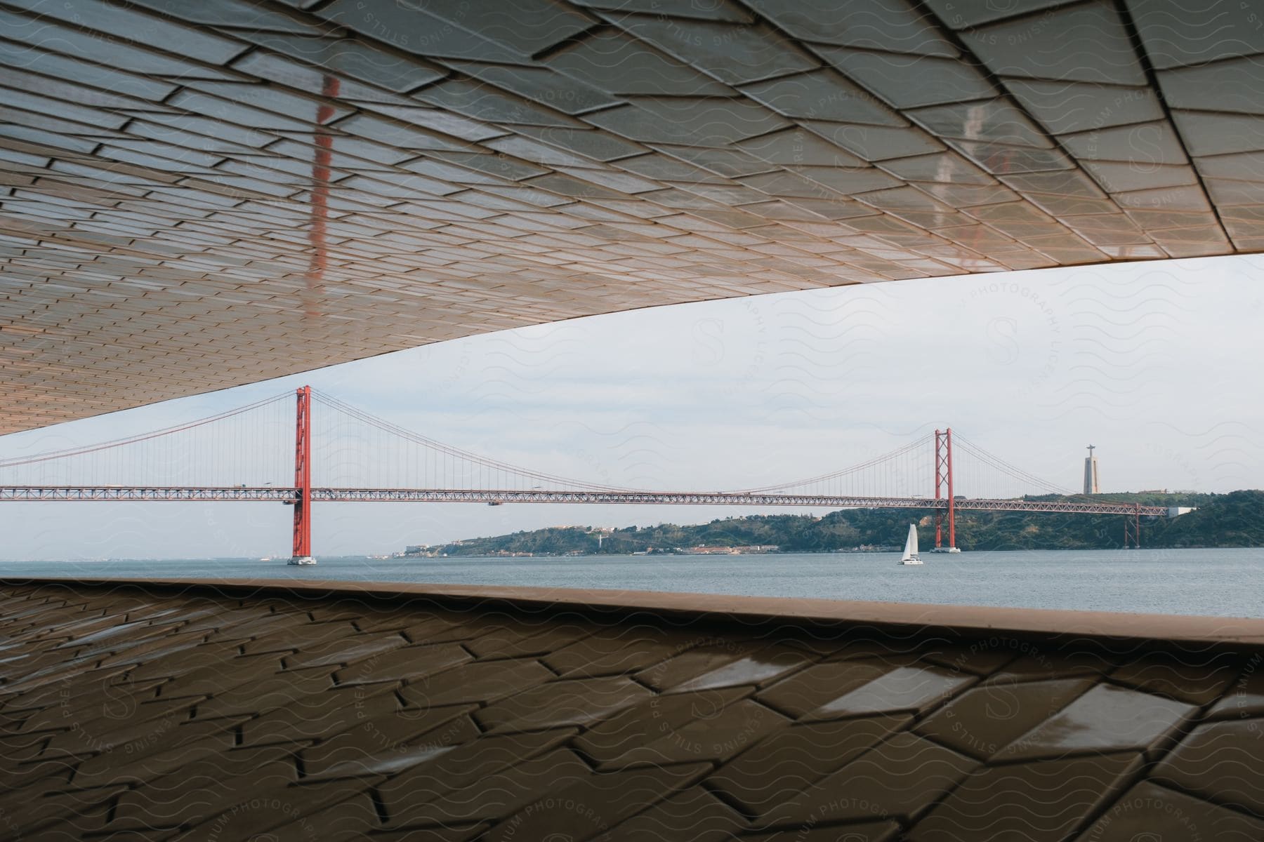 Inside a structure with a textured geometric surface as a sailboat is in the water near a suspension bridge stretching to the coast