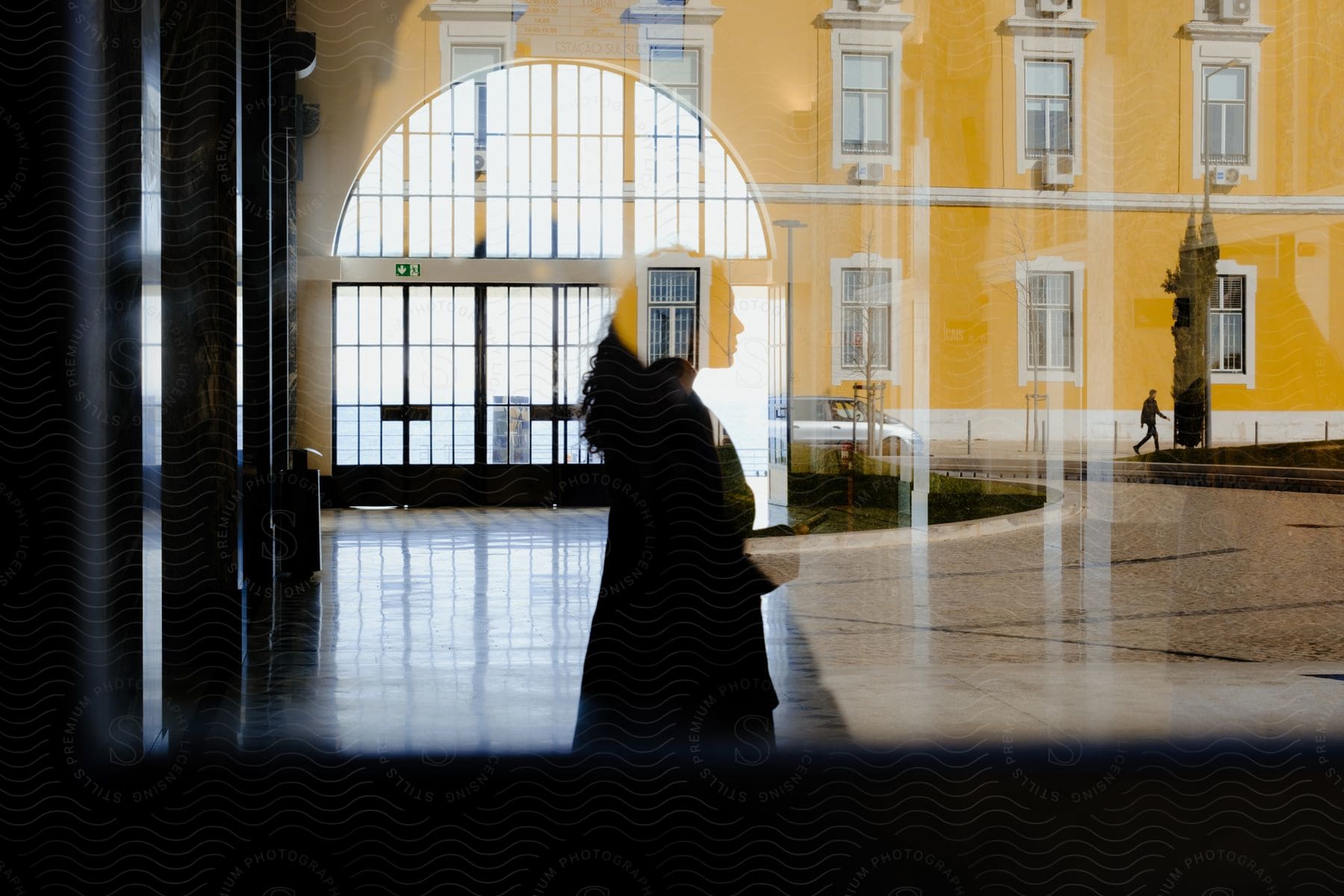 The reflection of a woman's silhouette in front of an arched entryway layered over a bright yellow building.