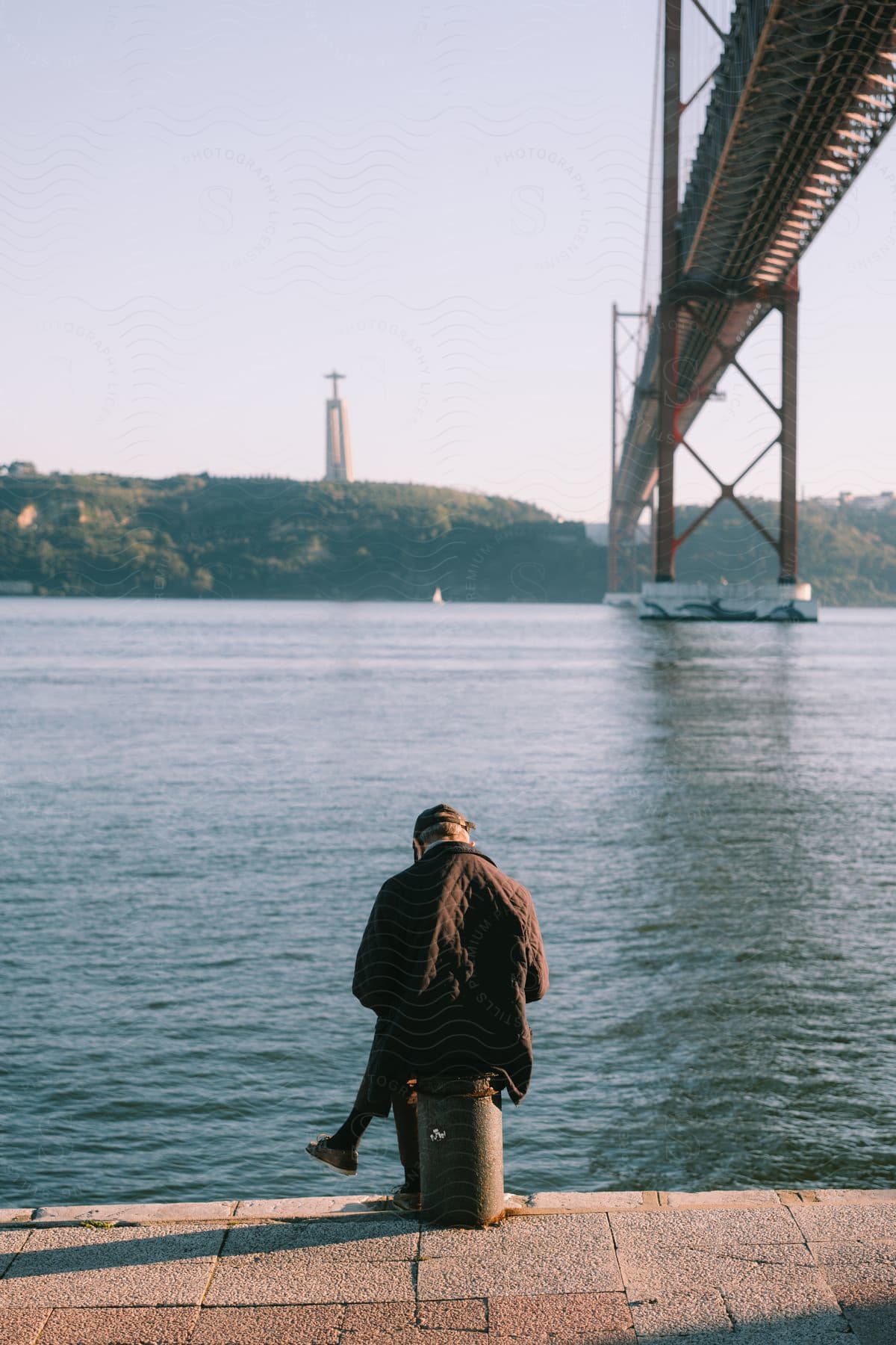 A person sits on the river shore, close to the flowing river. To the right, a steel train bridge stands tall, while on the other side of the river, lush green vegetation and a tower form a beautiful backdrop to this serene setting.