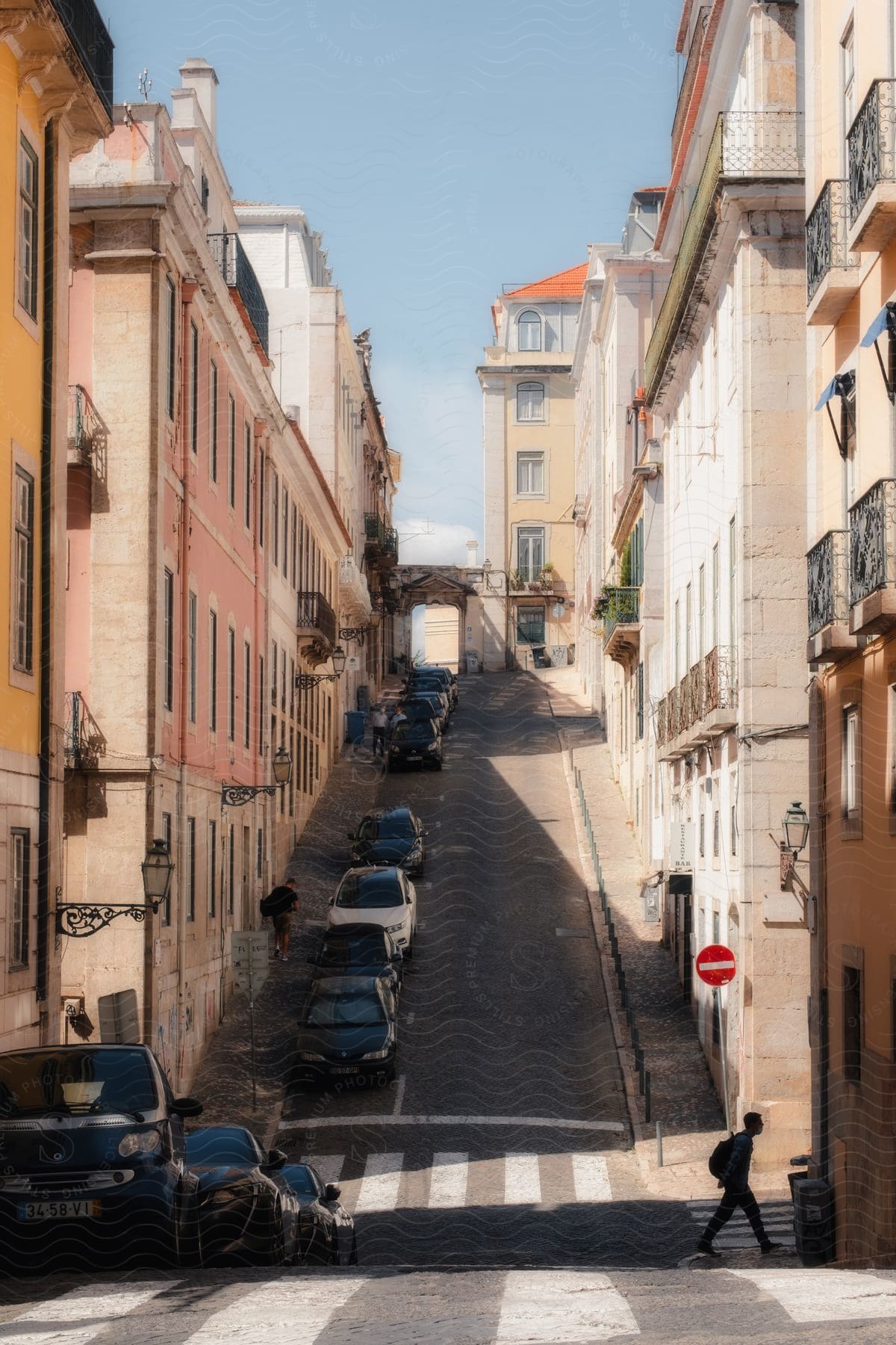 A man, carrying a backpack, walks across a street built on a hill. The road is lined with cars parked beside apartment buildings, creating a shaded area for the man while the sun shines brightly.