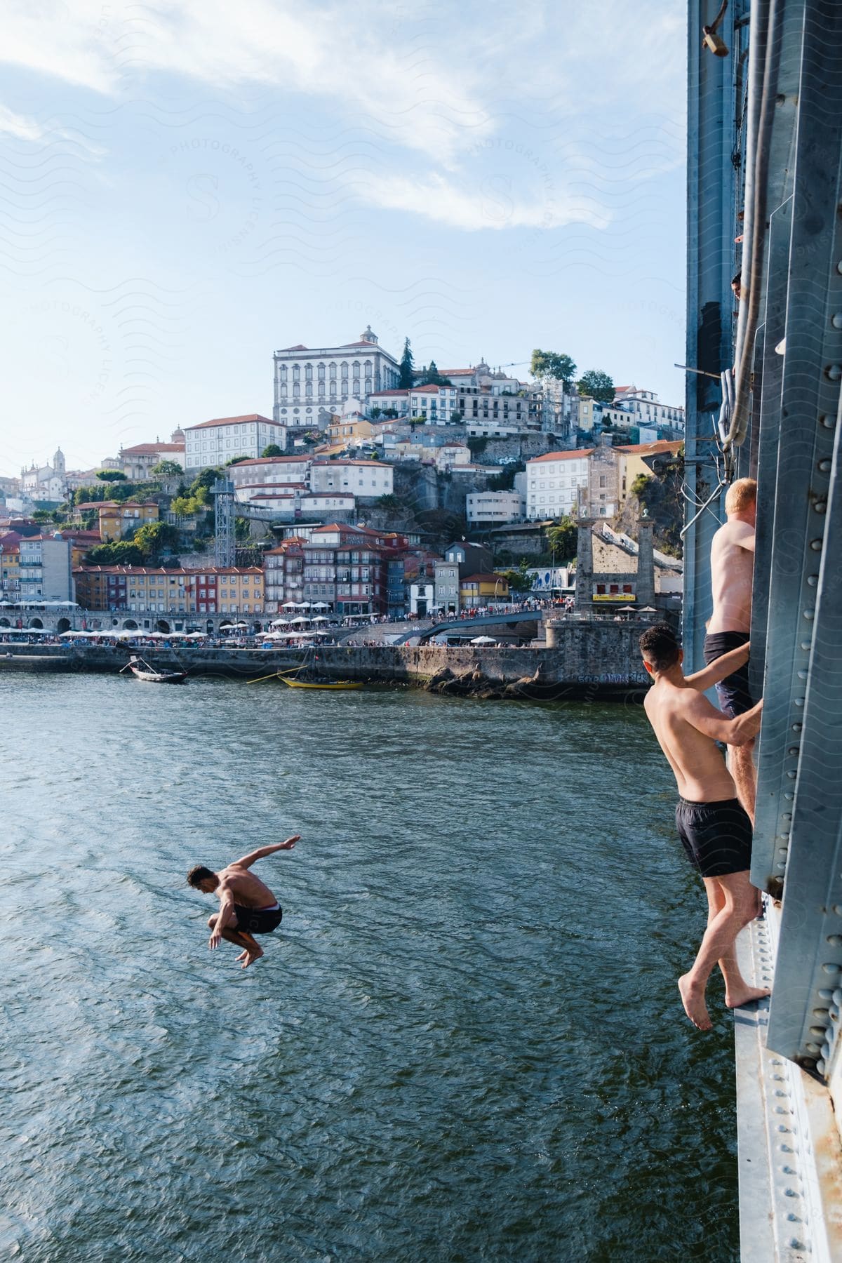 Three men wearing shorts, two holding onto a steel bridge while standing on the edge, and one jumping into the water in mid-air. In the background, there is a city view with two canoe docked at the shore