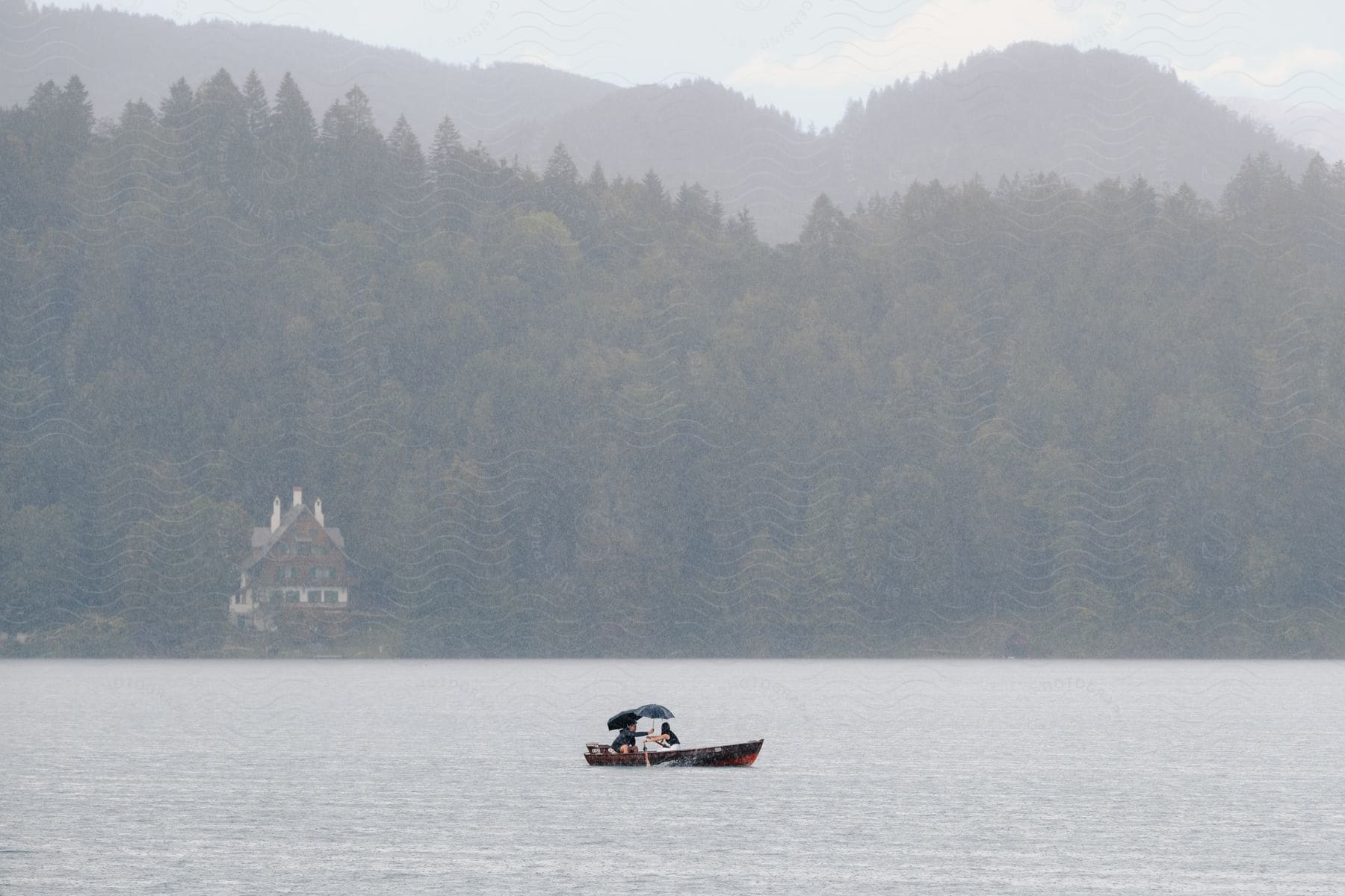 A man and woman are caught in the rain while in a row boat on a lake with forested hills in the background in the daytime.