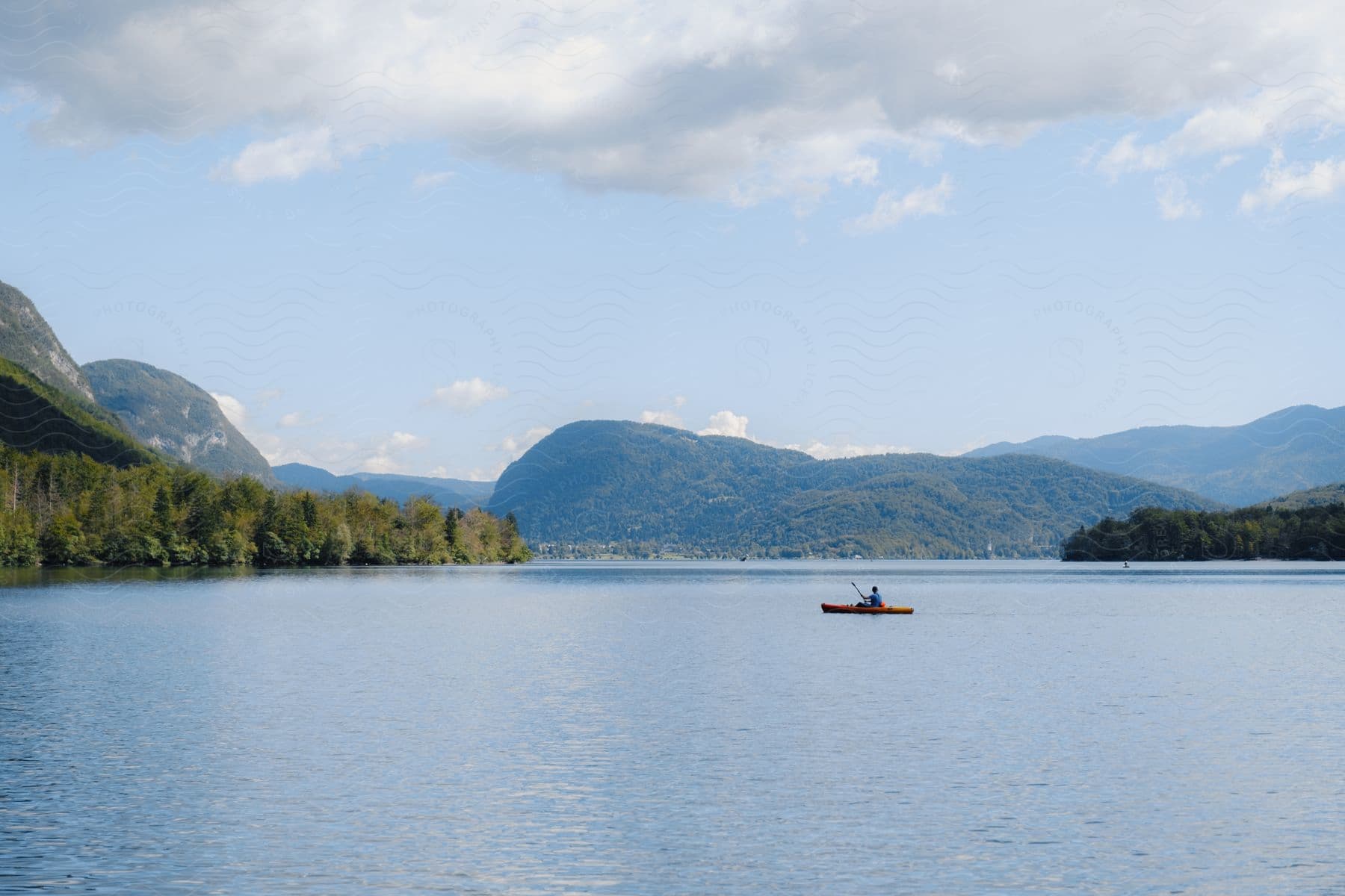 Person sits in boat in middle of lake surrounded by forest and hills on cloudy day.