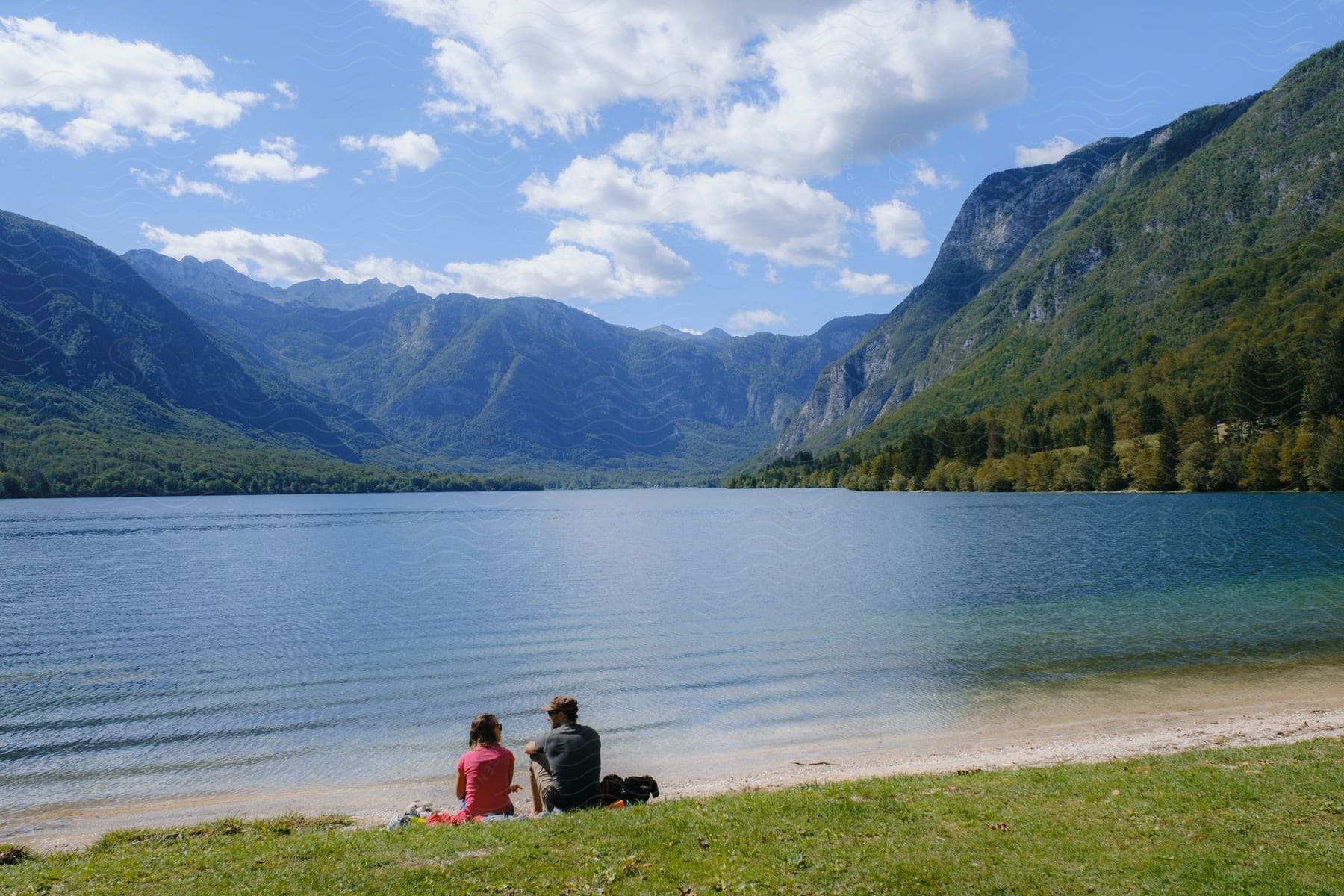 A couple looking at each other on the shore of a lake and in the background several mountains full of trees and vegetation.