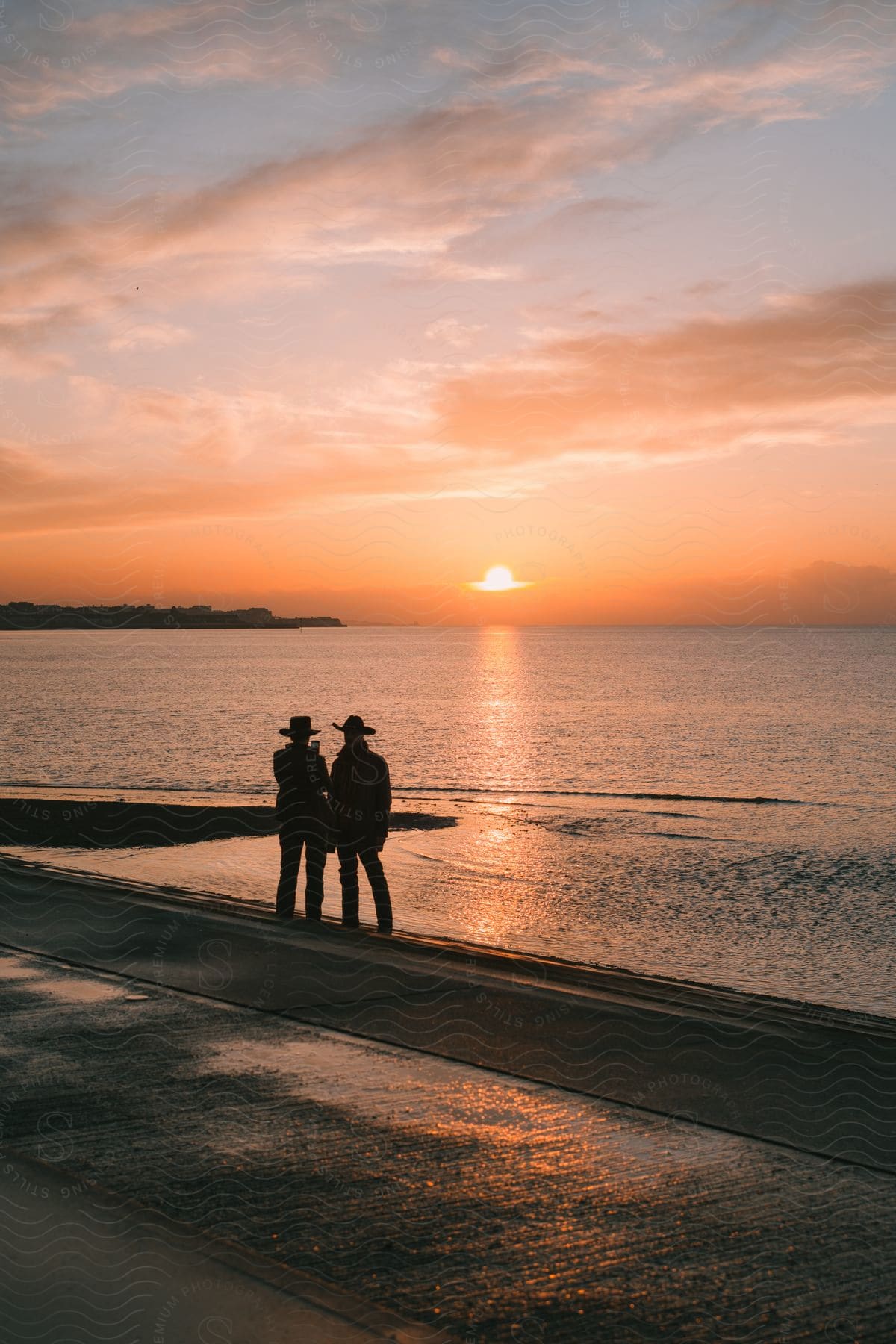 Silhouettes of two people wearing cowboy hats standing in the sand looking out at the sun setting on the ocean.