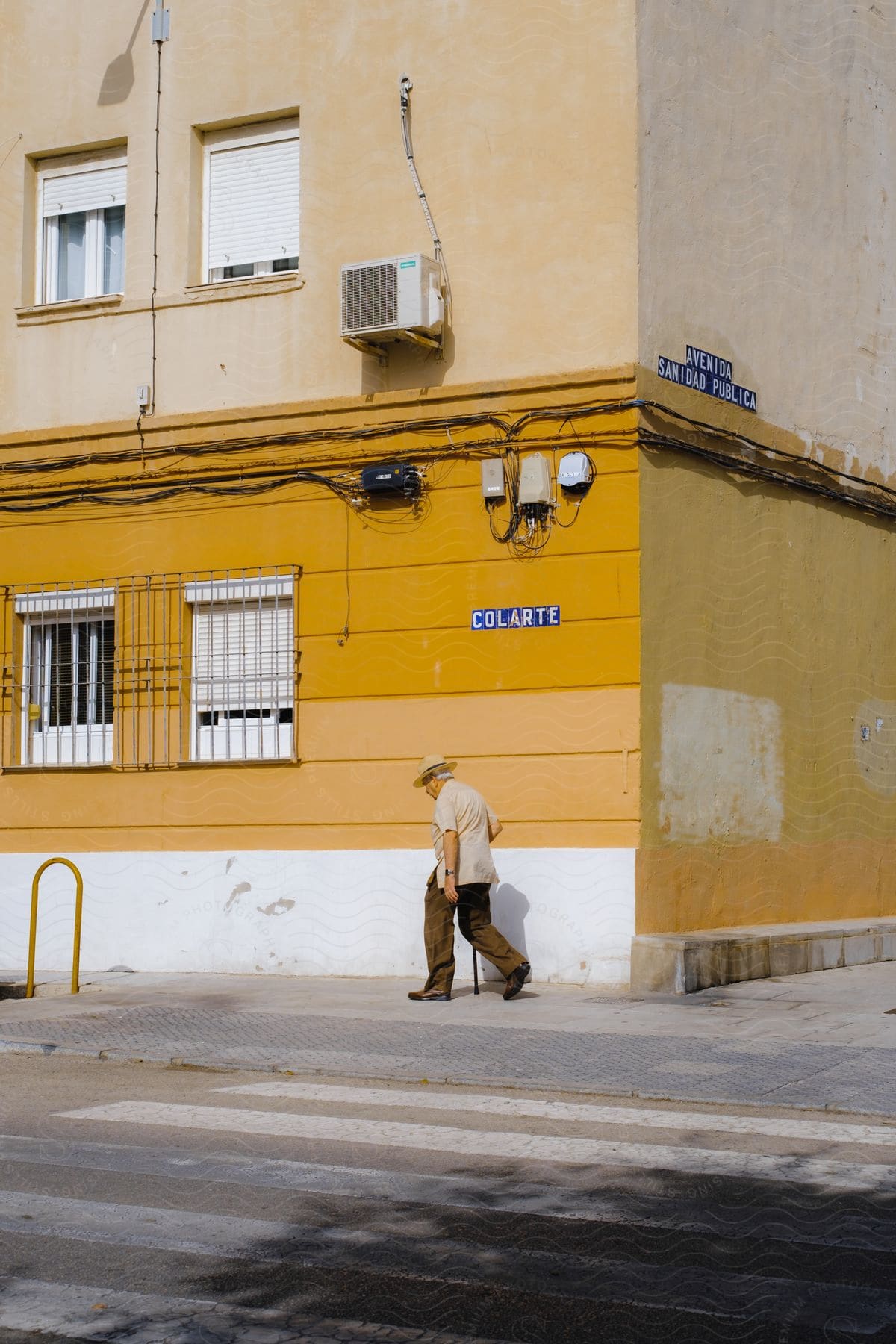 An old man using a cane walking down a street next to a yellow wall.