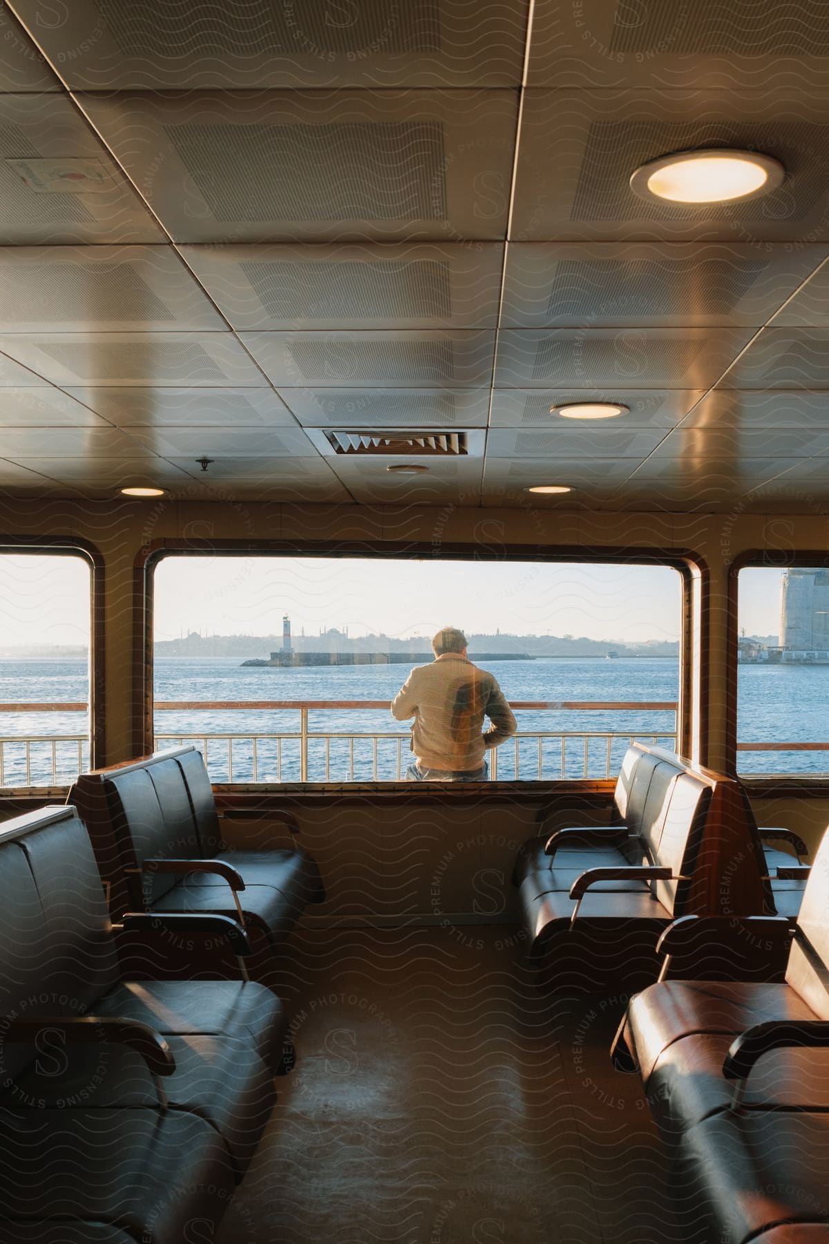 The inside of building or ship with seats lining the windows overlooking water, as a man stand outside with his back to the window.