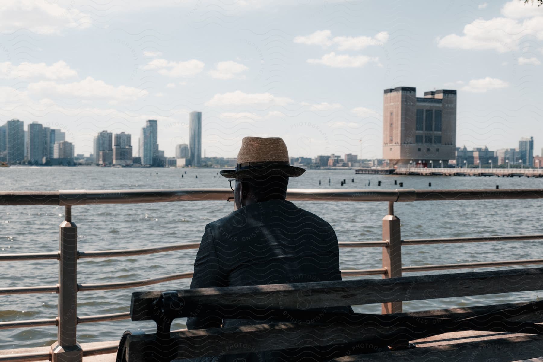 A man is sitting on a bench near a body of water enjoying the view of the city buildings.