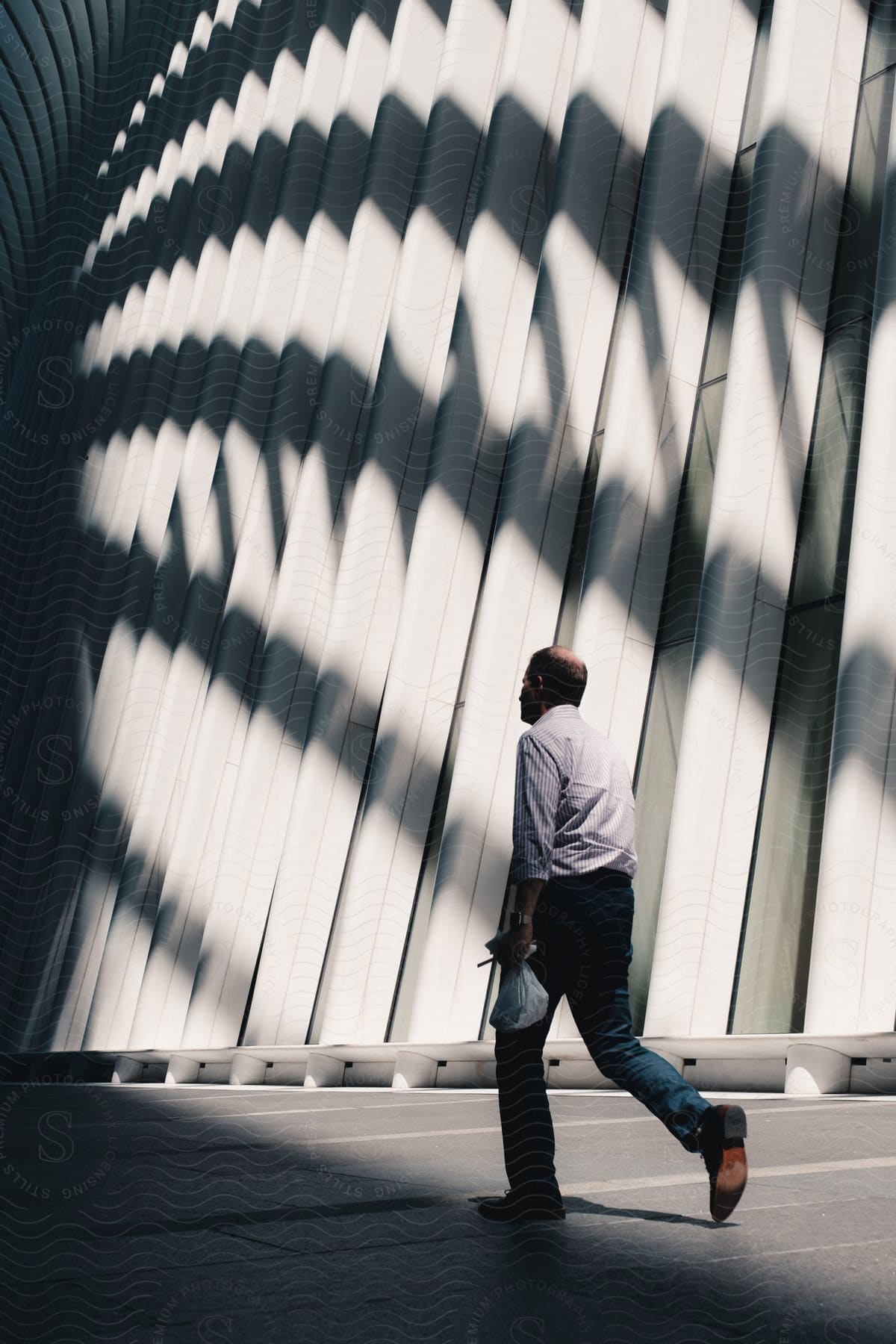 A man carrying a plastic bag is walking outside in a city near a building with modern architecture and interesting shadows being cast in the daytime.