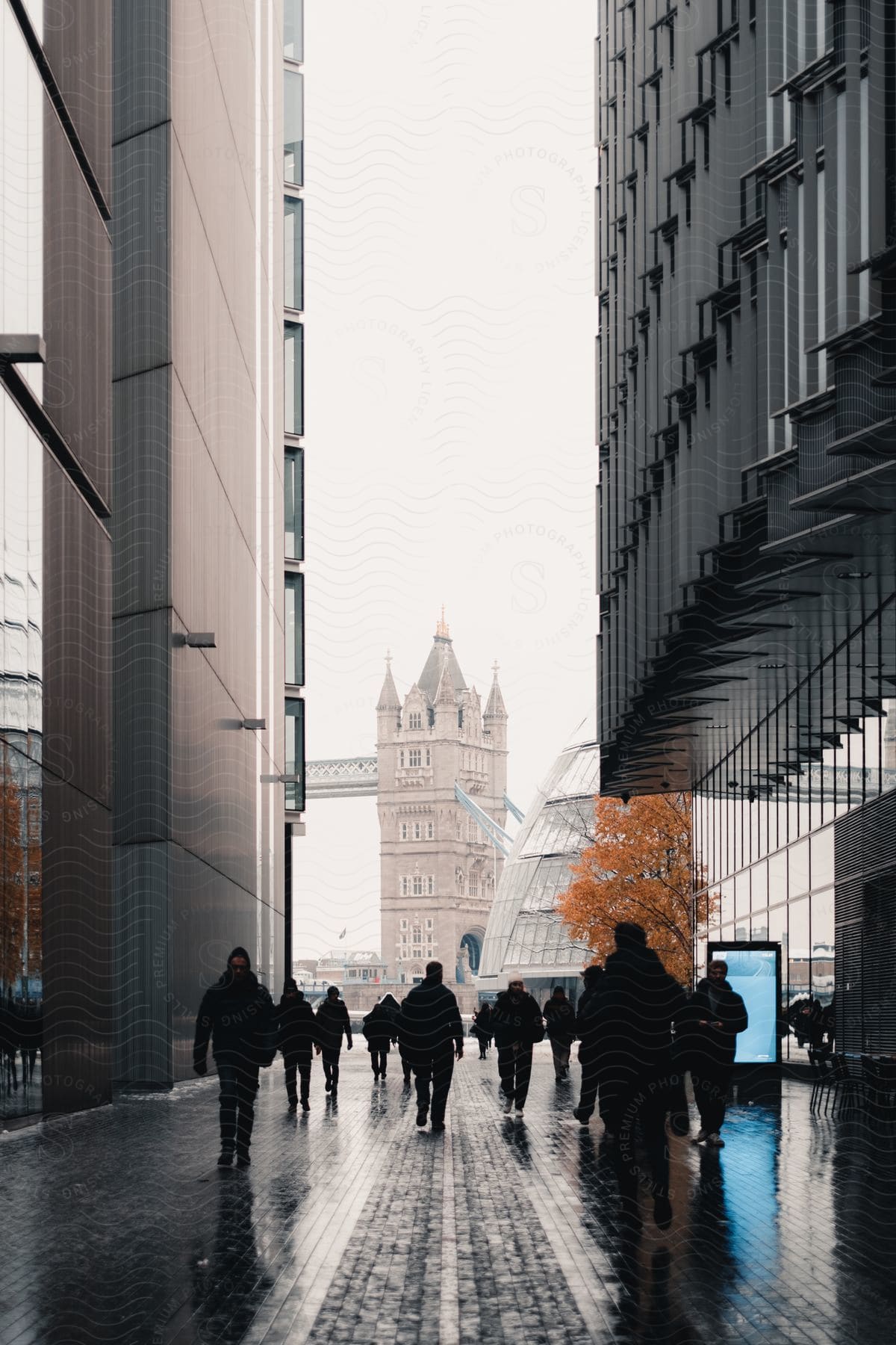 Pedestrians are walking through an outdoor corridor between two modern buildings with a castle-like tower in the background on an overcast day.