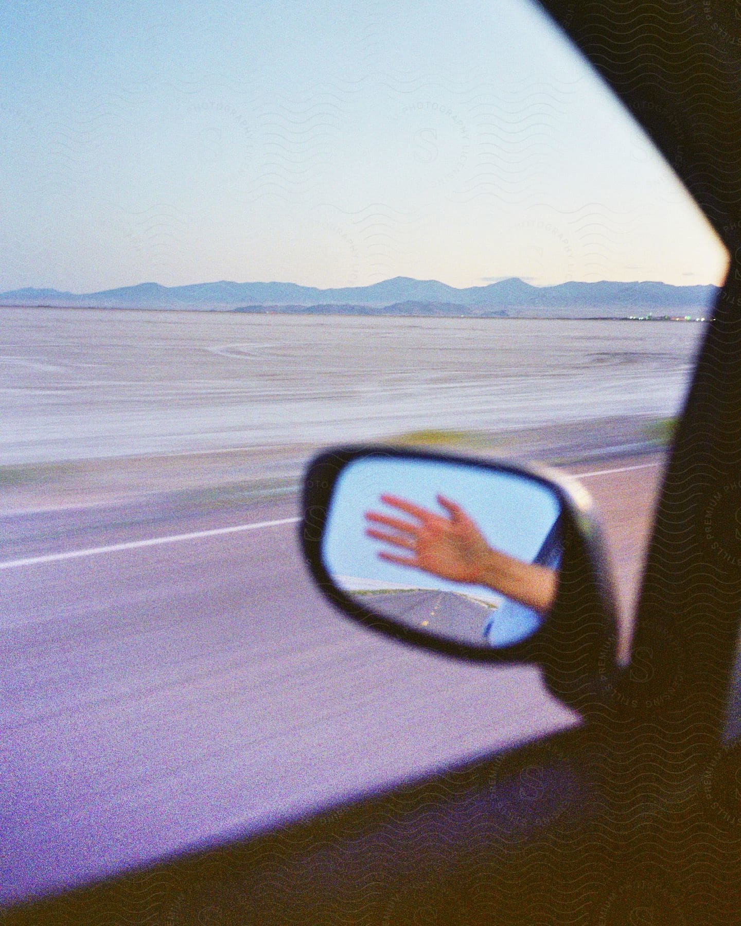 Side mirror reflects hand of person on road trip past sandy plain near mountains.