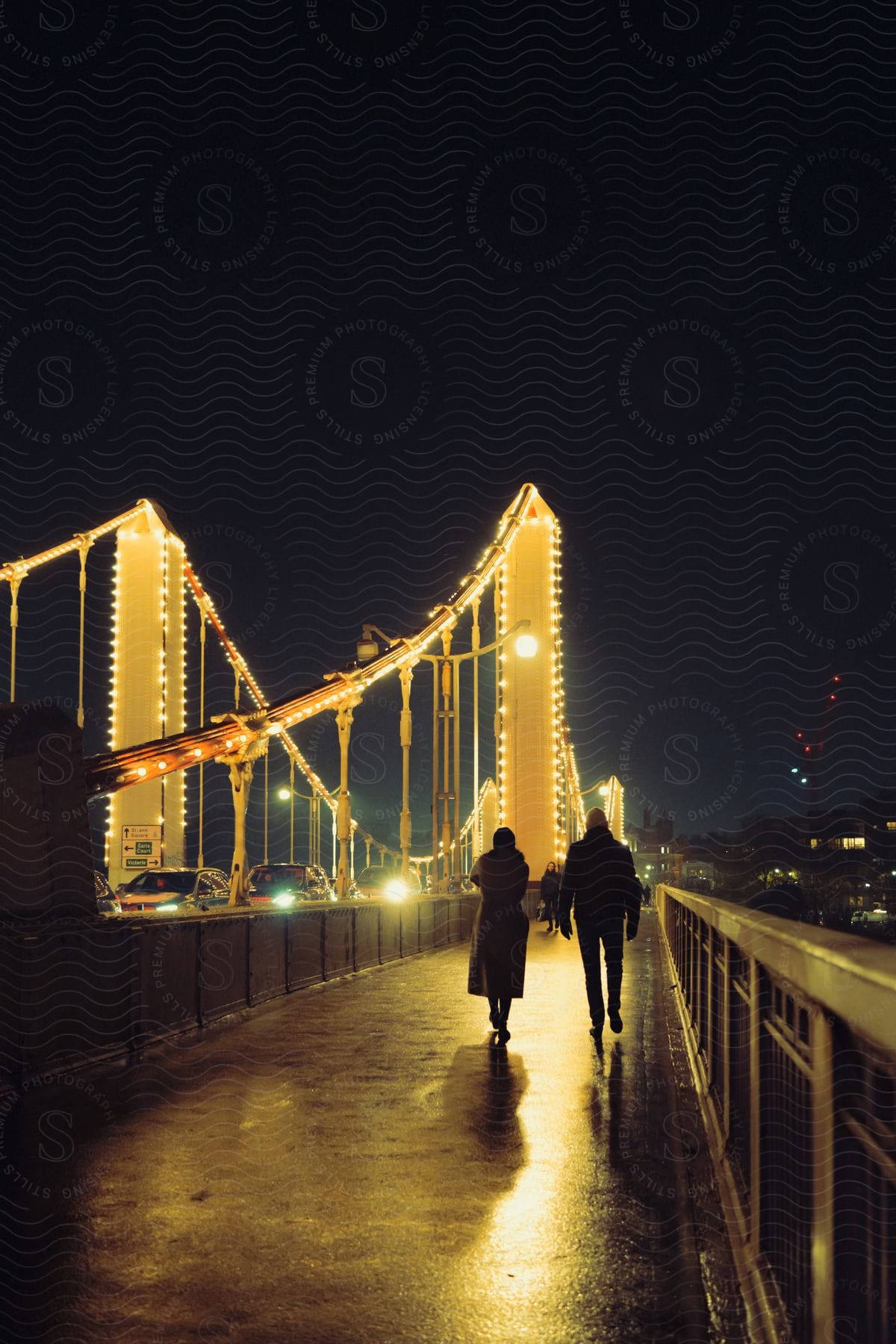 In the cold night, two people walk on a well-lit bridge walkpath, illuminated by a good lighting system. Cars drive by on the bridge, and in the backdrop, the city view adds to the scene.