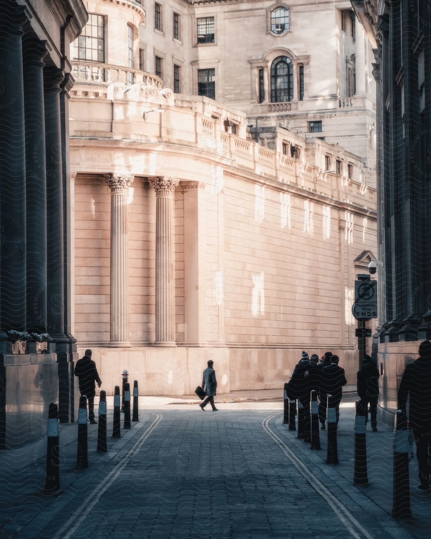 People walking across the street and on the sidewalk of a street with stone pavement, in front of a classically designed building.