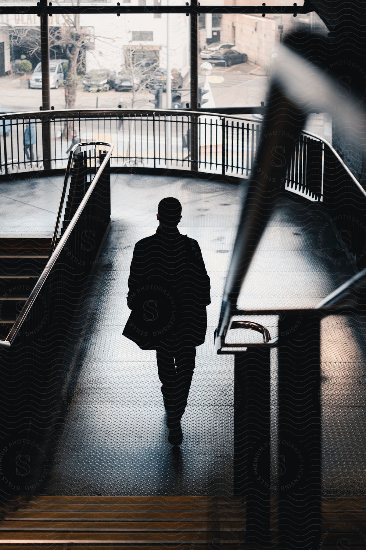 A man is walking alone on a dimly lit landing toward another flight of stairs in a building with large windows in the background during the day.