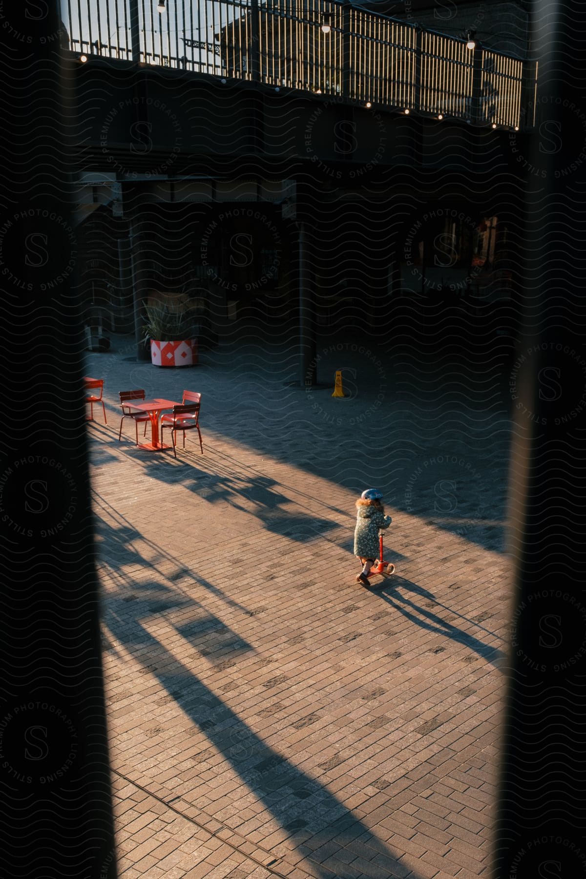 Child riding a scooter in a courtyard with red tables.