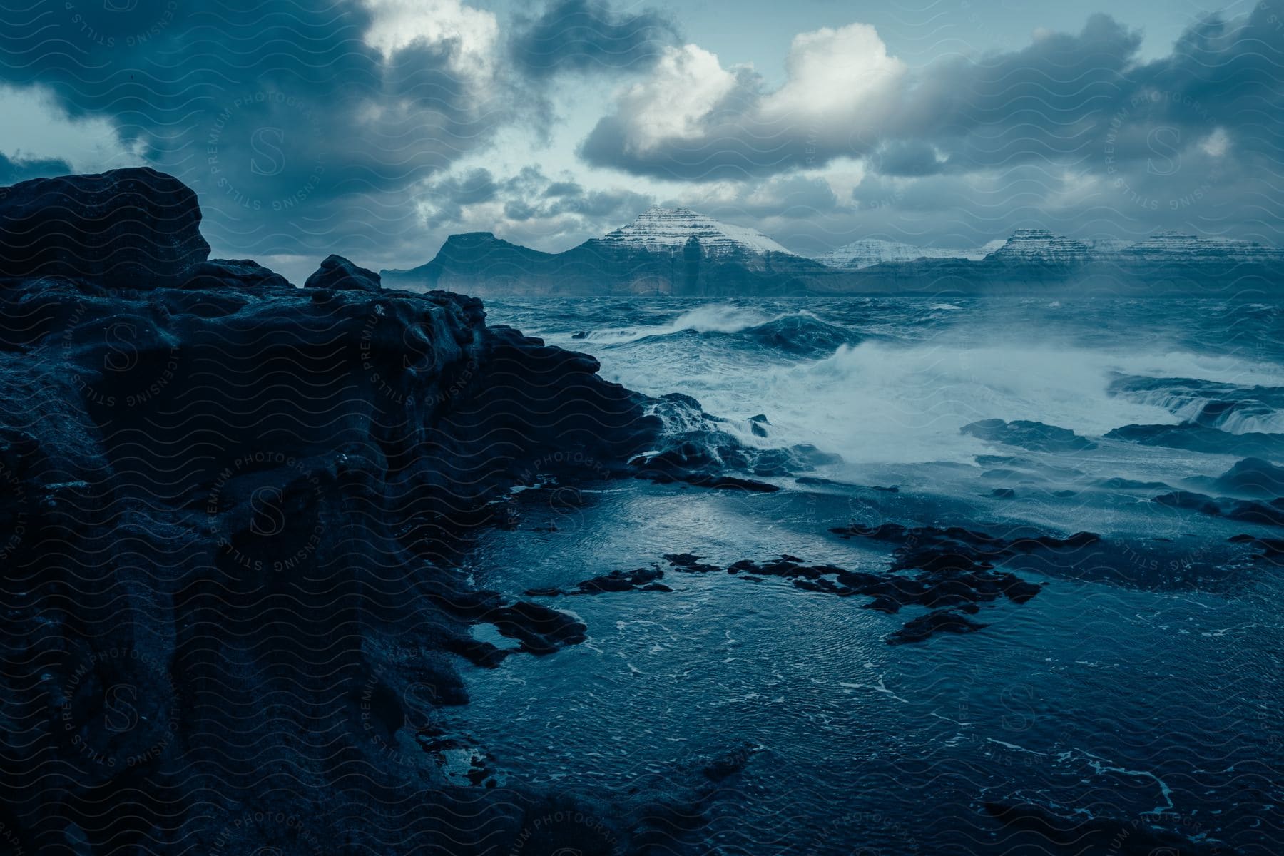 cloudy landscape of a rocky shore with powerful waves crashing against the rocks, mountains in the distance.