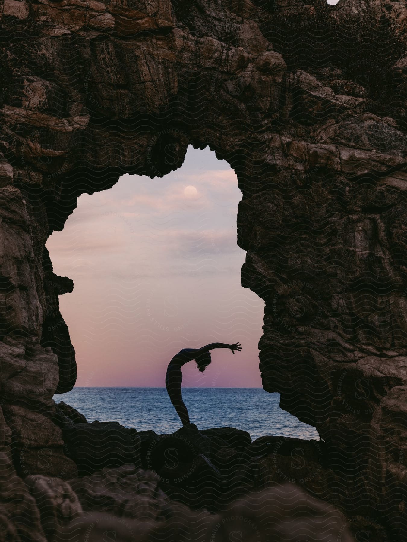 Silhouette of a woman posing with her arms behind her on top of a rocky cave in the middle of the ocean.